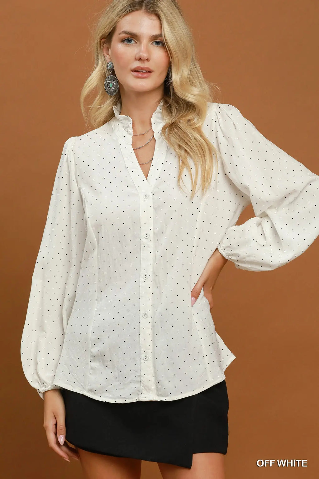 A woman modeling a long-sleeve off-white button-up blouse with a small black polka dot pattern, paired with a black skirt, against a solid brown studio background.