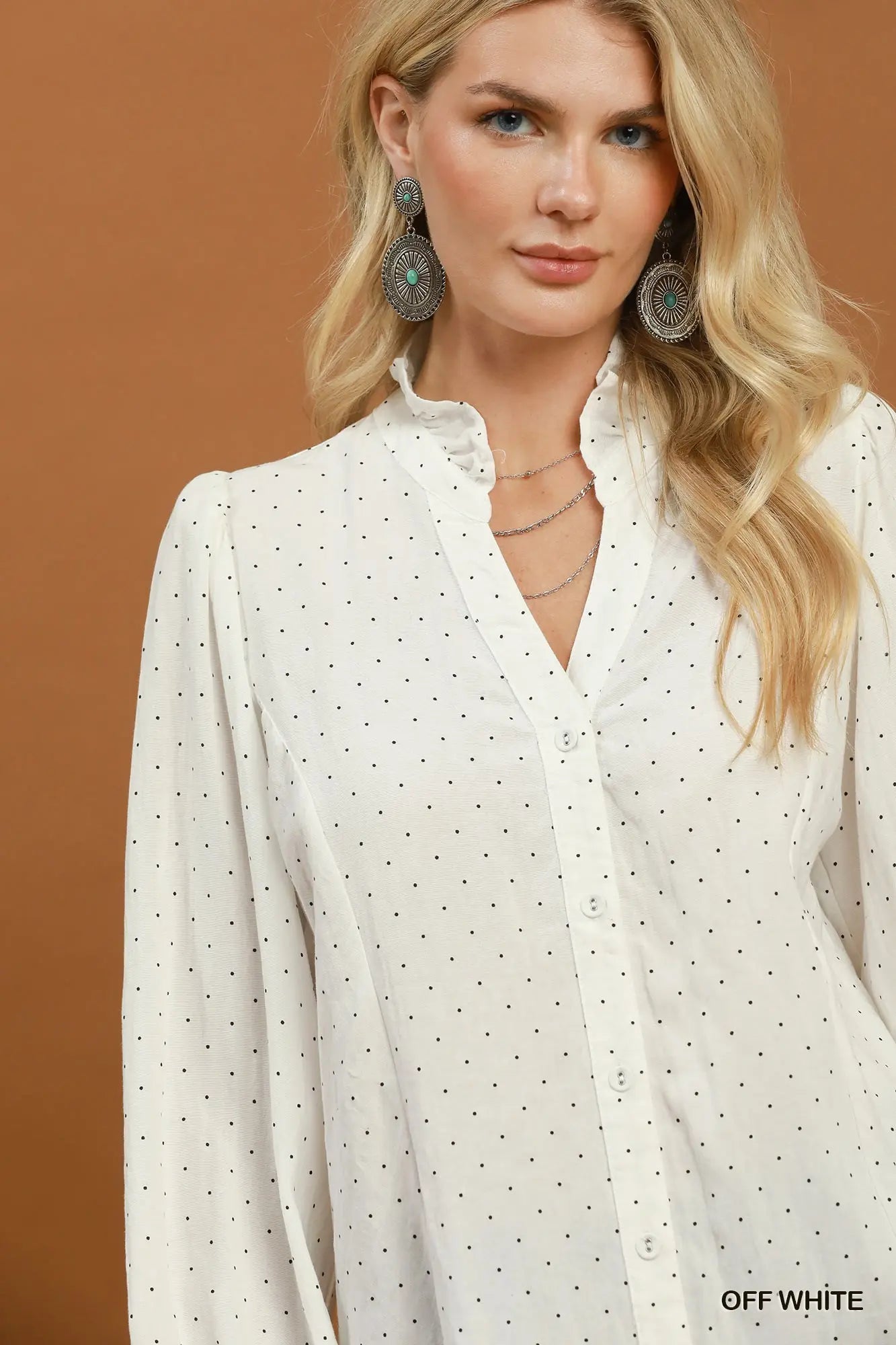 A woman wearing an off-white long-sleeve button-up blouse with a small black polka dot pattern, styled with layered silver necklaces and large circular earrings, set against a solid warm brown background.