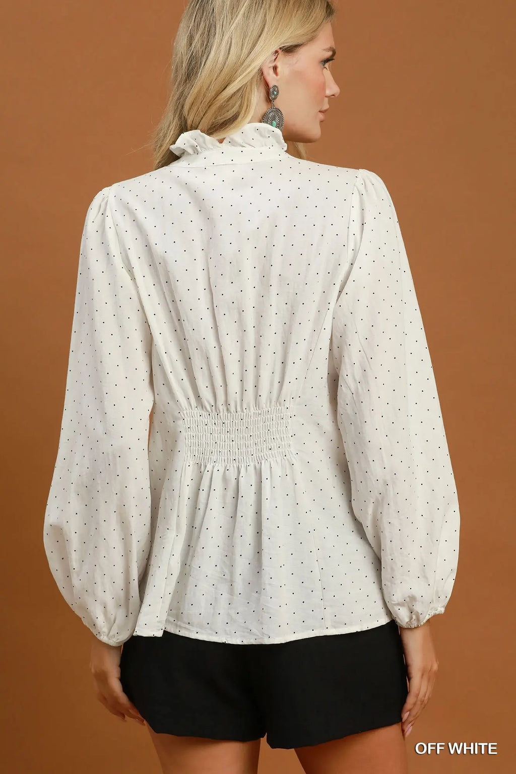 A woman shown from the back wearing an off-white long-sleeve blouse with tiny black polka dots, a ruffled high neck, and a smocked waist, paired with black shorts against a solid brown studio background.
