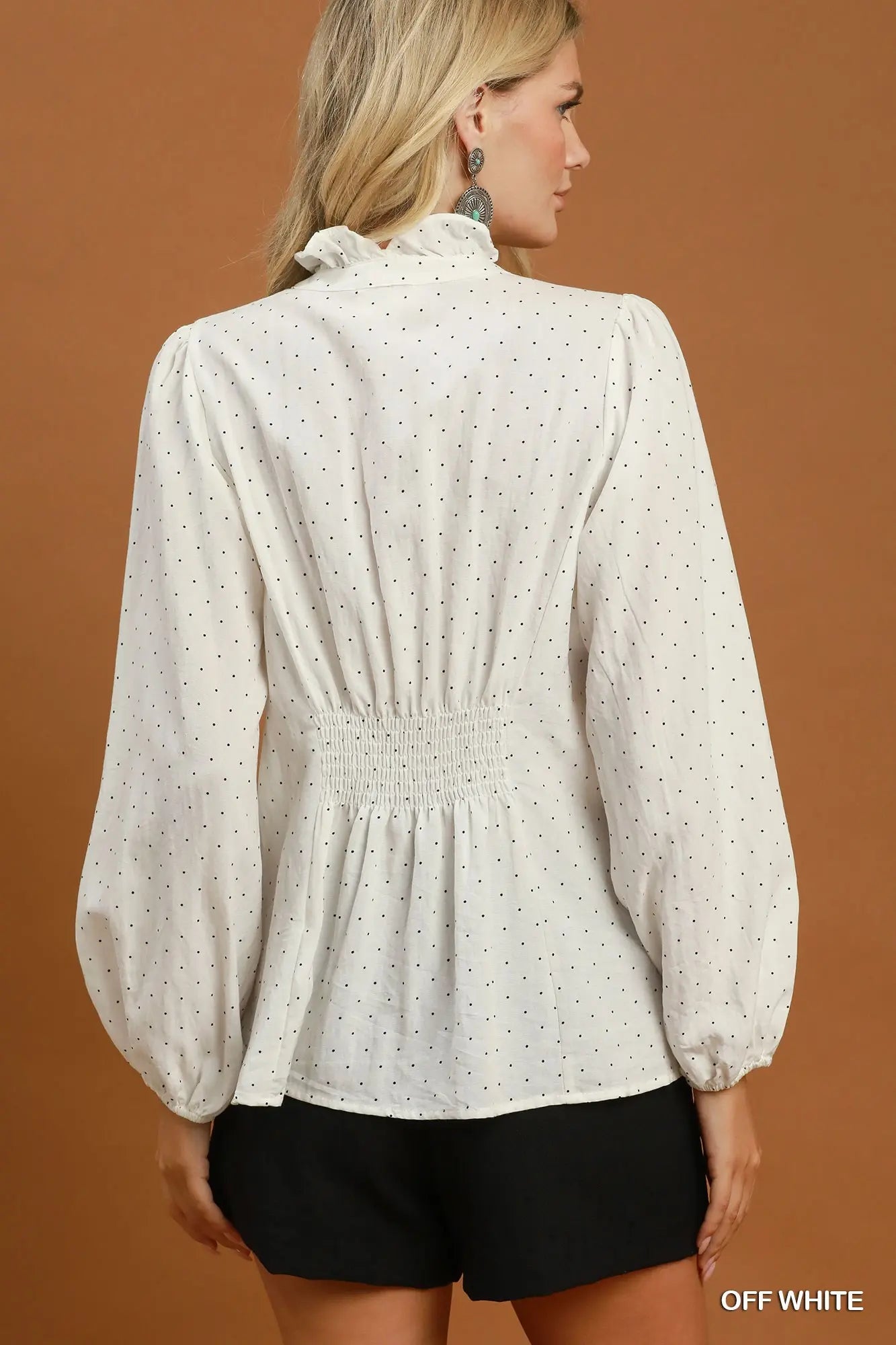 A woman shown from the back wearing an off-white long-sleeve blouse with tiny black polka dots, a ruffled high neck, and a smocked waist, paired with black shorts against a solid brown studio background.