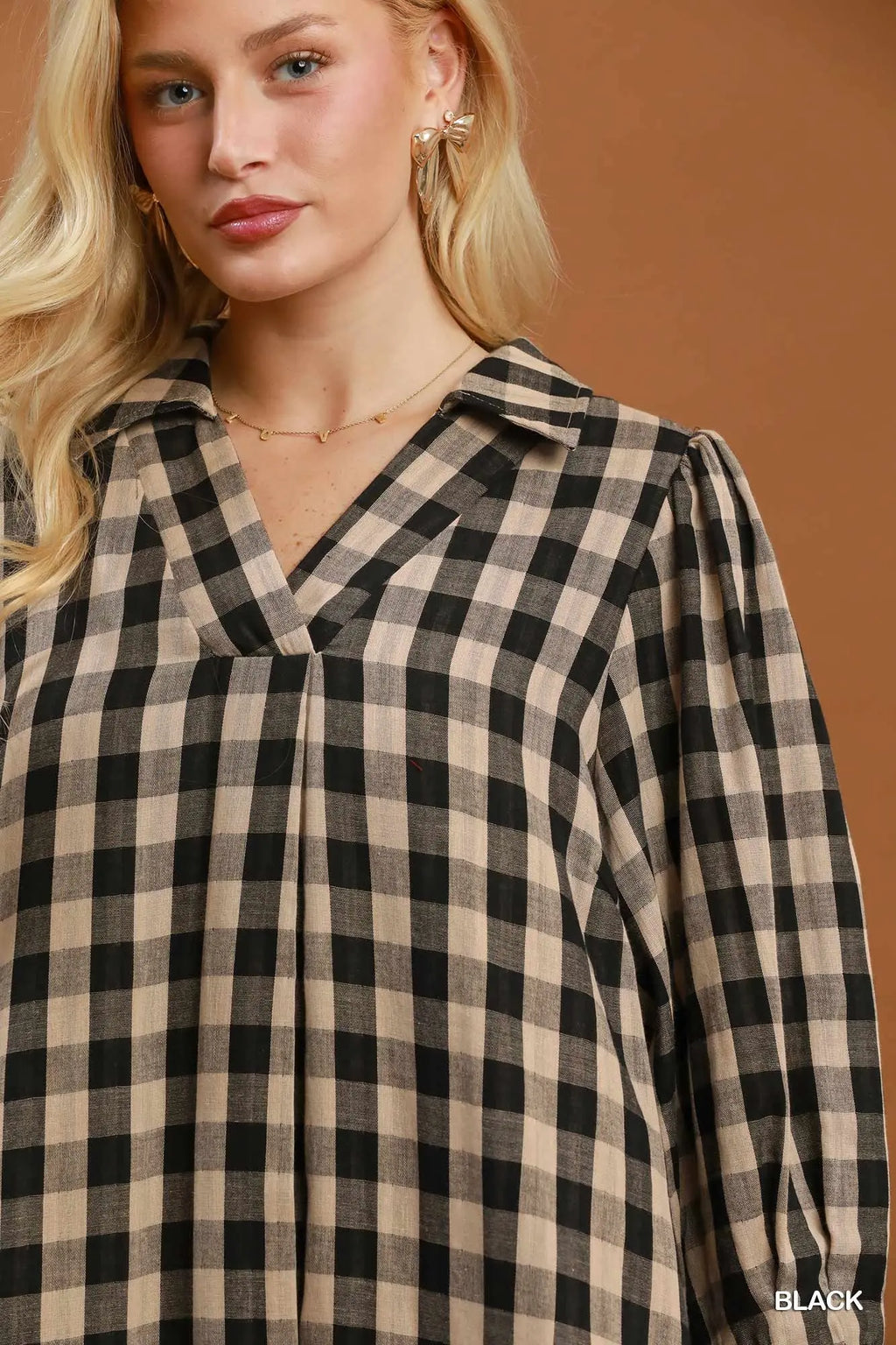 A close-up of a model wearing a black and beige gingham plaid long-sleeve blouse with a V-neckline, set against a solid warm brown background.