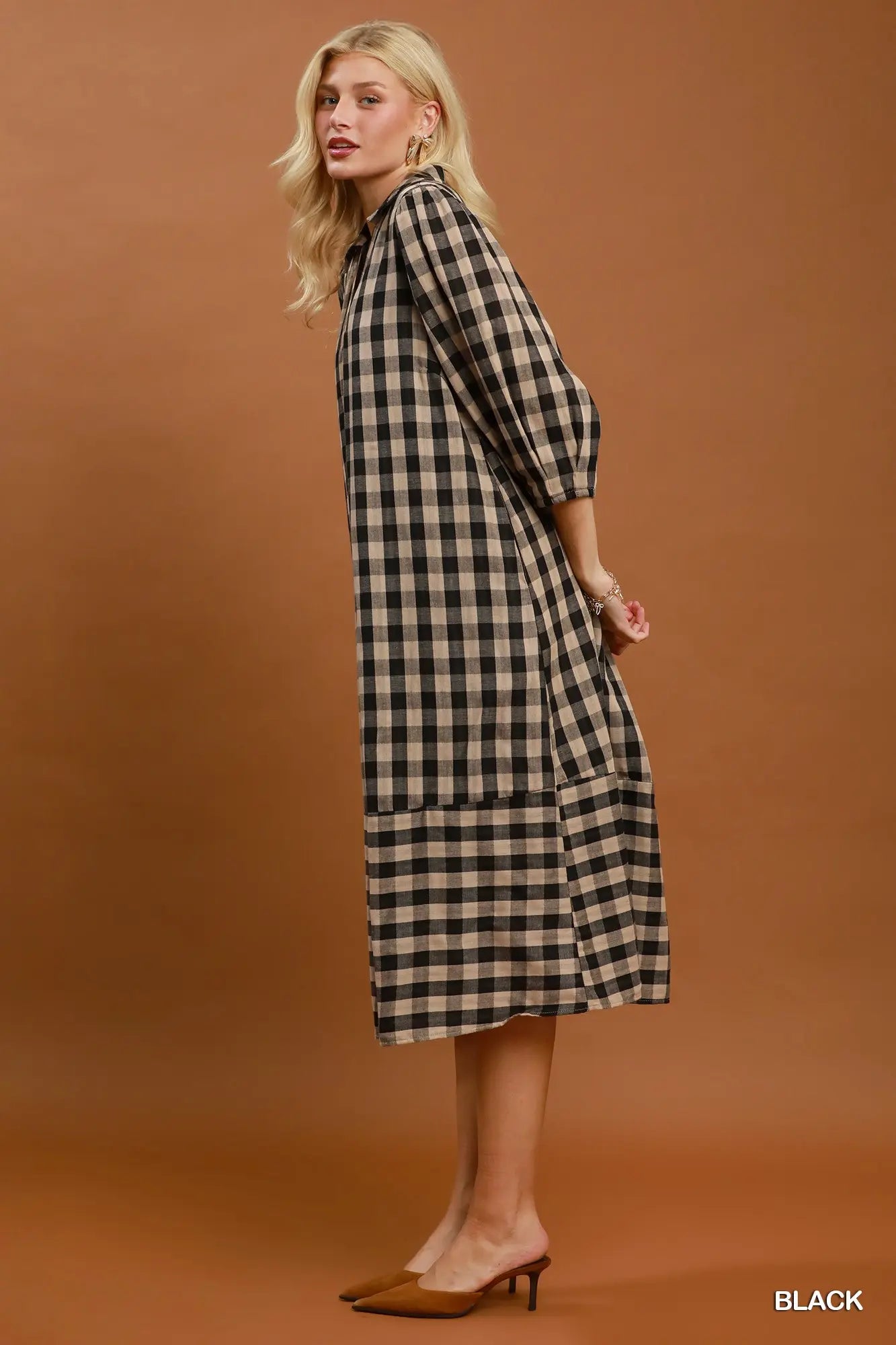 A woman modeling a black and beige gingham midi dress with three-quarter sleeves, standing against a solid warm brown studio background. She is wearing tan pointed-toe mule heels, and the word 'BLACK' is printed in the bottom right corner.