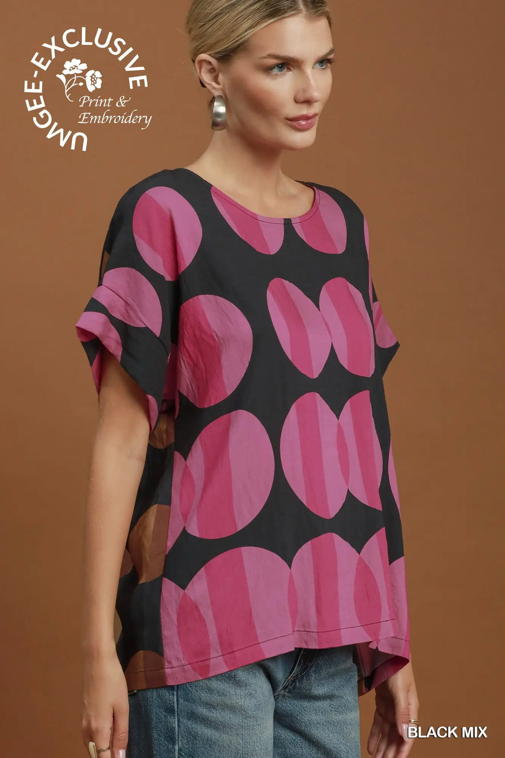 A woman wearing a black short-sleeve top with a bold pink and magenta abstract circular pattern, paired with blue jeans, set against a solid brown studio background. A white circular logo for 'Ungee Exclusive Print & Embroidery' is visible in the top left, and 'BLACK MIX' is written in the bottom right corner.