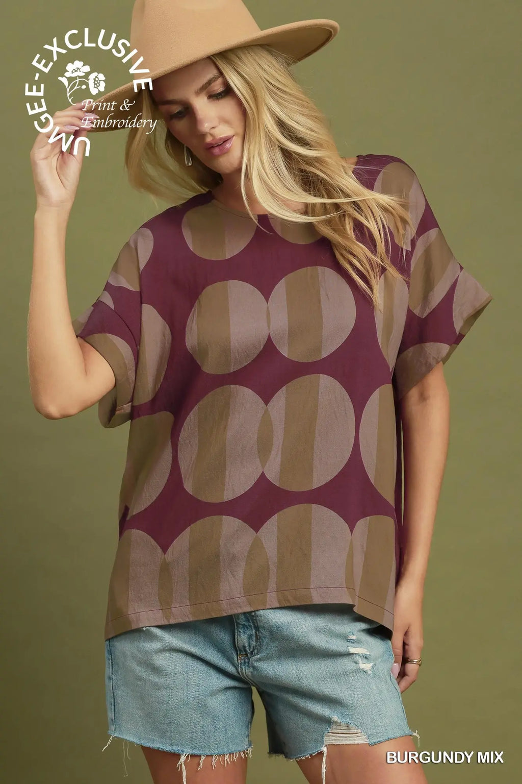 A woman wearing a burgundy and tan oversized short-sleeve top with a large circular pattern, paired with light-wash denim shorts and a tan hat, against an olive green background. A 'GEE-EXCLUSIVE' watermark is visible on the top left.