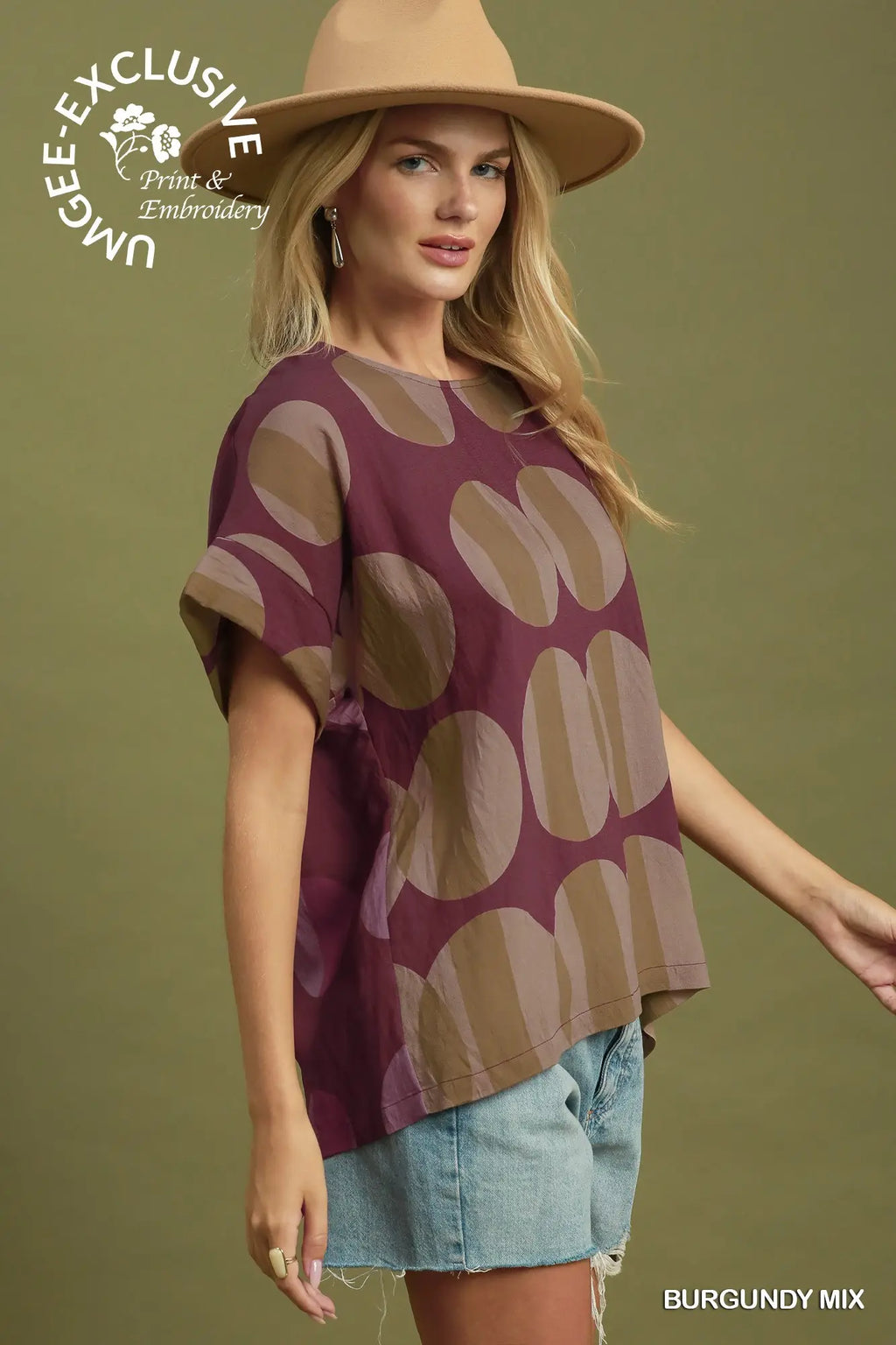 A woman modeling a short-sleeve burgundy top with a large beige oval pattern, paired with denim shorts and a tan hat, against a solid olive green background. A logo for 'Ungee Exclusive Print & Embroidery' is visible in the top left, and 'BURGUNDY MIX' is written in the bottom right.