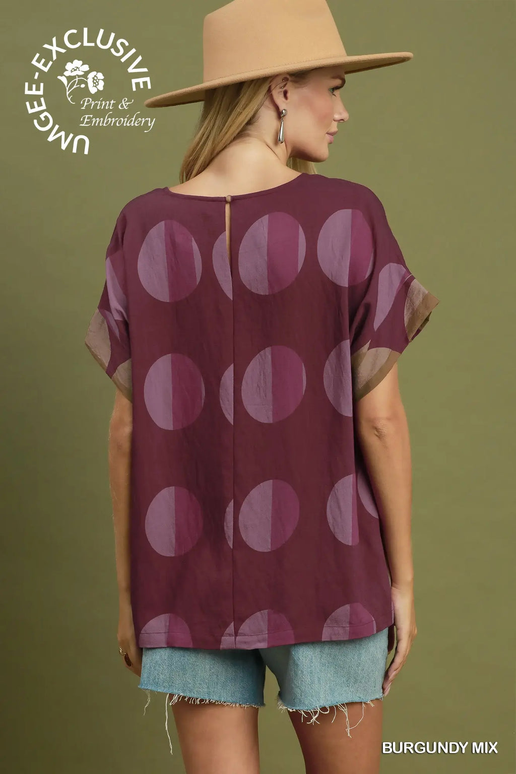 A woman shown from the back wearing a burgundy short-sleeve top with a large tonal polka dot pattern, paired with denim shorts and a tan hat, against an olive green background. A logo for 'Ungee Exclusive Print & Embroidery' is visible in the top left, and 'BURGUNDY MIX' is written in the bottom right.