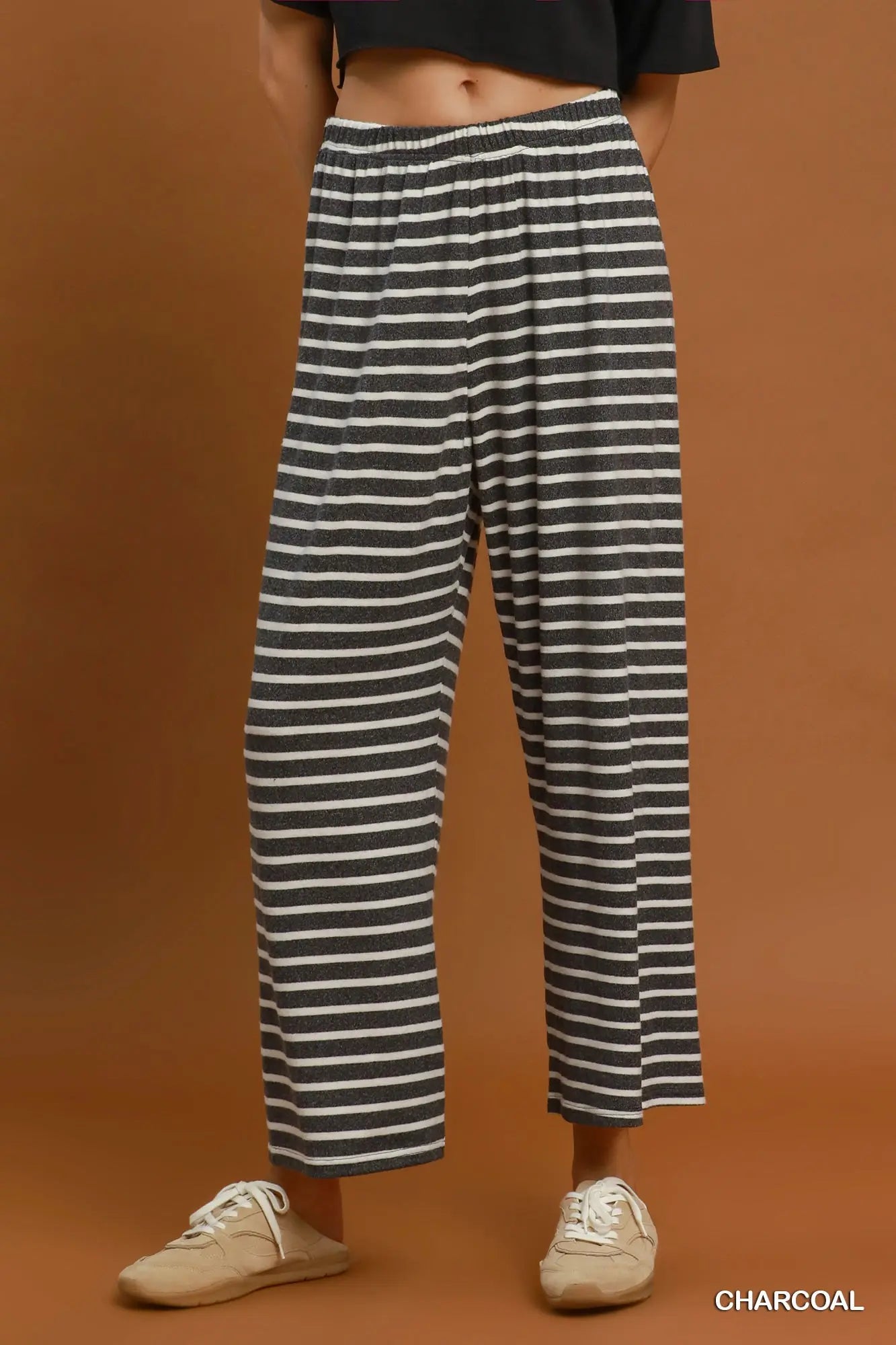 A person wearing loose charcoal and off-white horizontally striped wide-leg cropped pants, paired with a black cropped top and beige sneakers, against a solid warm brown studio background. The word 'CHARCOAL' appears in the bottom right corner.