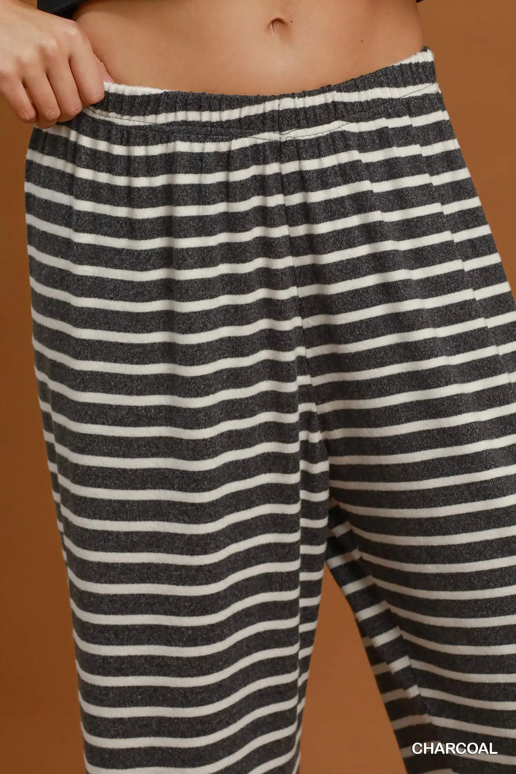 Close-up of a person wearing striped lounge pants in charcoal and off-white, with the hand pulling the elastic waistband. The word 'CHARCOAL' appears in the bottom right corner.