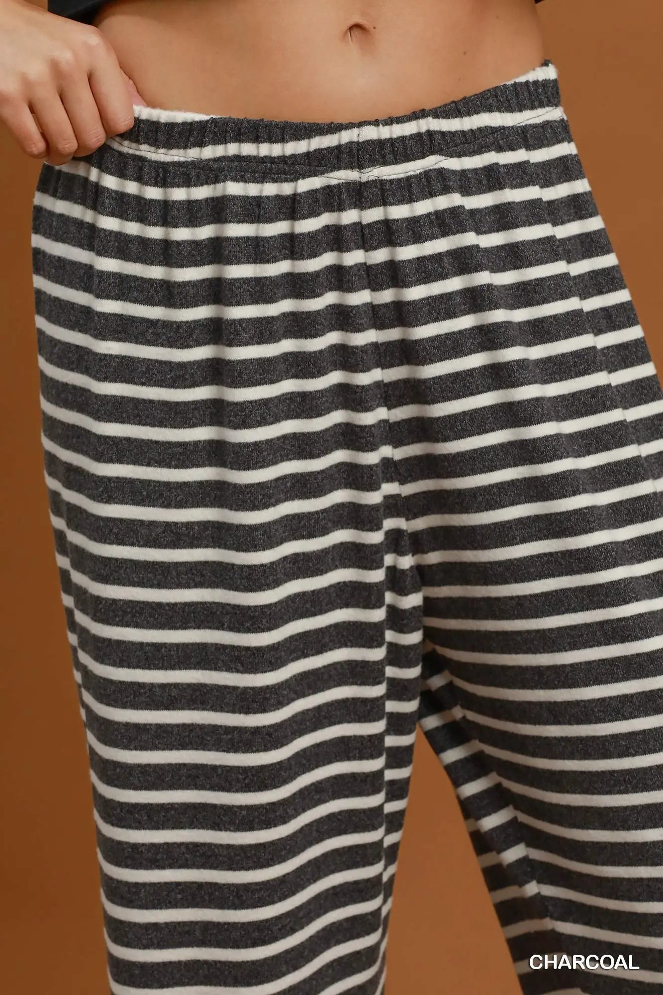 Close-up of a person wearing striped lounge pants in charcoal and off-white, with the hand pulling the elastic waistband. The word 'CHARCOAL' appears in the bottom right corner.