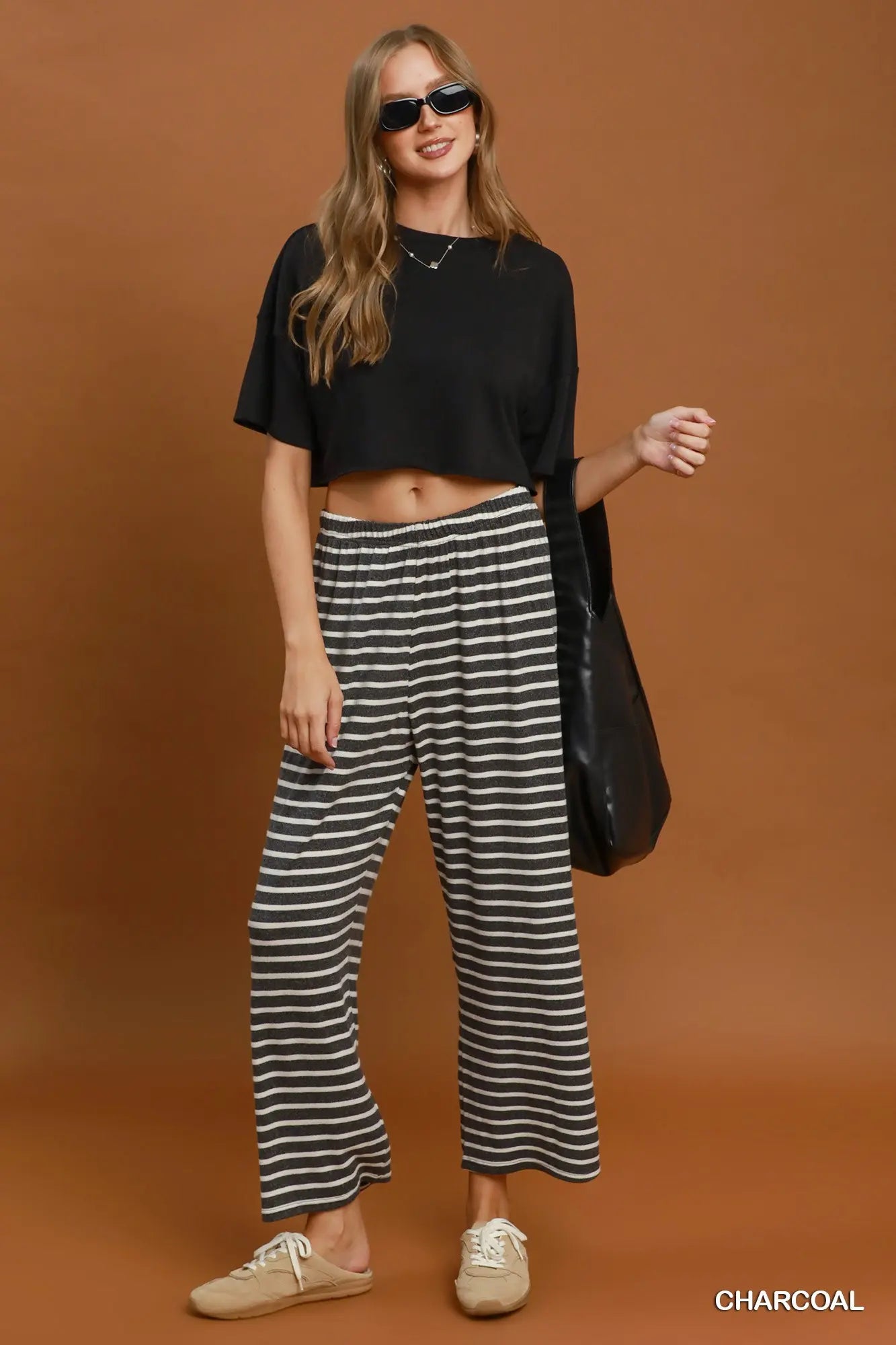 A woman modeling a black cropped t-shirt and charcoal gray and black striped wide-leg pants, styled with beige slip-on shoes and a black shoulder bag against a solid brown studio background.