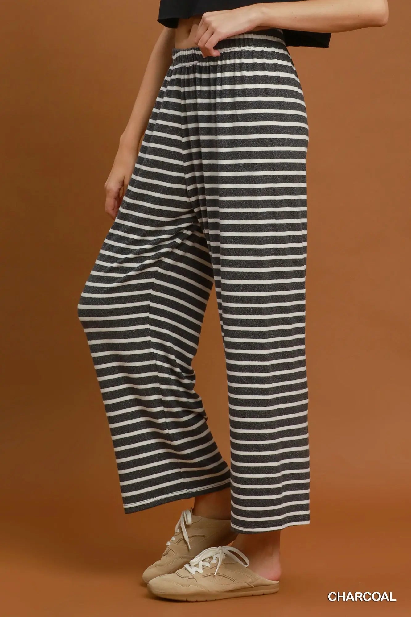 A person wearing loose charcoal and off-white horizontally striped wide-leg cropped pants, paired with beige slip-on shoes, against a solid warm brown studio background. The word 'CHARCOAL' appears in the bottom right corner.