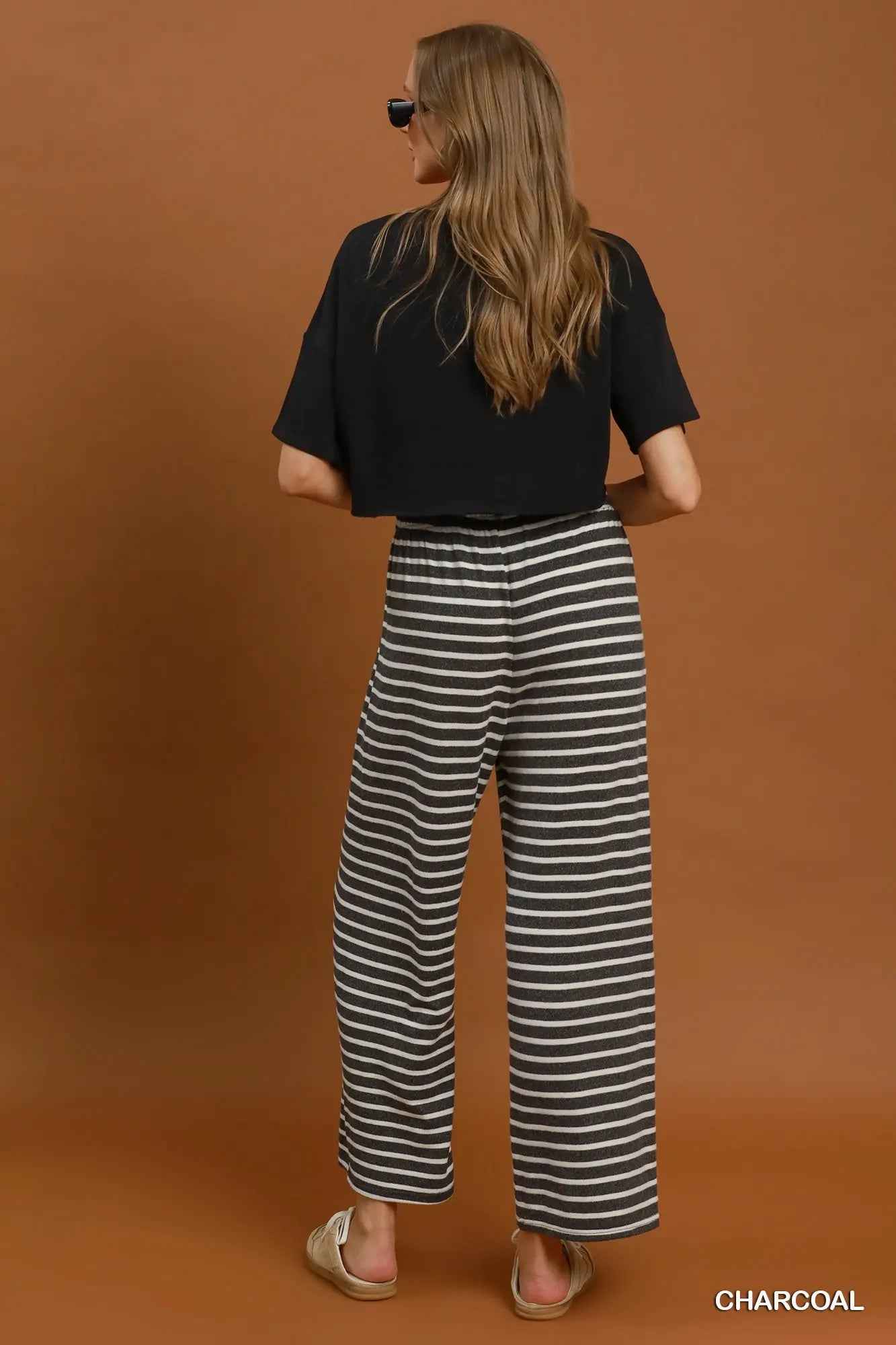 A woman stands with her back to the camera wearing charcoal wide-leg striped pants, a black short-sleeve top, and beige slide sandals against a solid brown studio backdrop. The text 'CHARCOAL' appears in the bottom right corner.