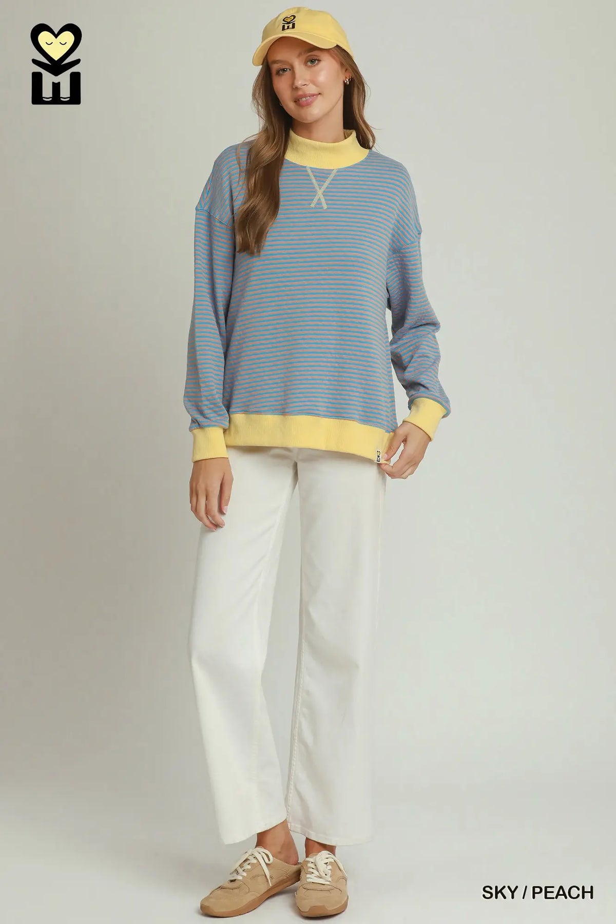 A woman modeling a light blue and peach striped long-sleeve knit sweater with yellow accents on the collar, cuffs, and hem, paired with white wide-leg pants, beige sneakers, and a yellow baseball cap against a light gray studio background.