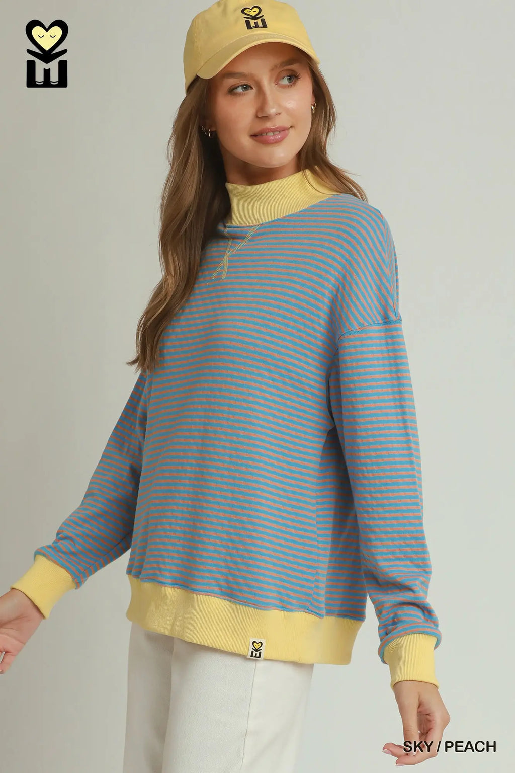 A woman wearing a light blue and peach-orange striped long-sleeve mock neck sweatshirt with yellow ribbed trim, paired with a yellow cap and white pants, against a light gray studio background.