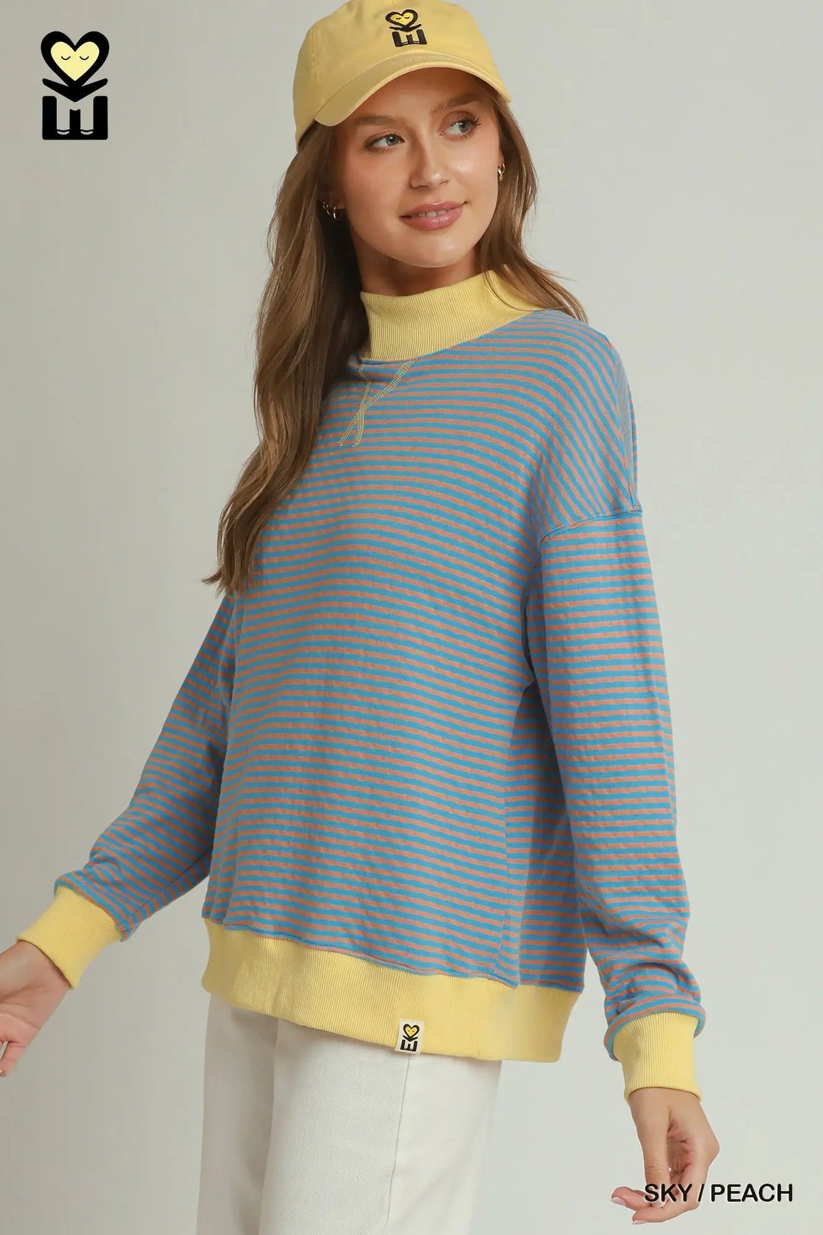 A woman wearing a light blue and peach-orange striped long-sleeve mock neck sweatshirt with yellow ribbed trim, paired with a yellow cap and white pants, against a light gray studio background.