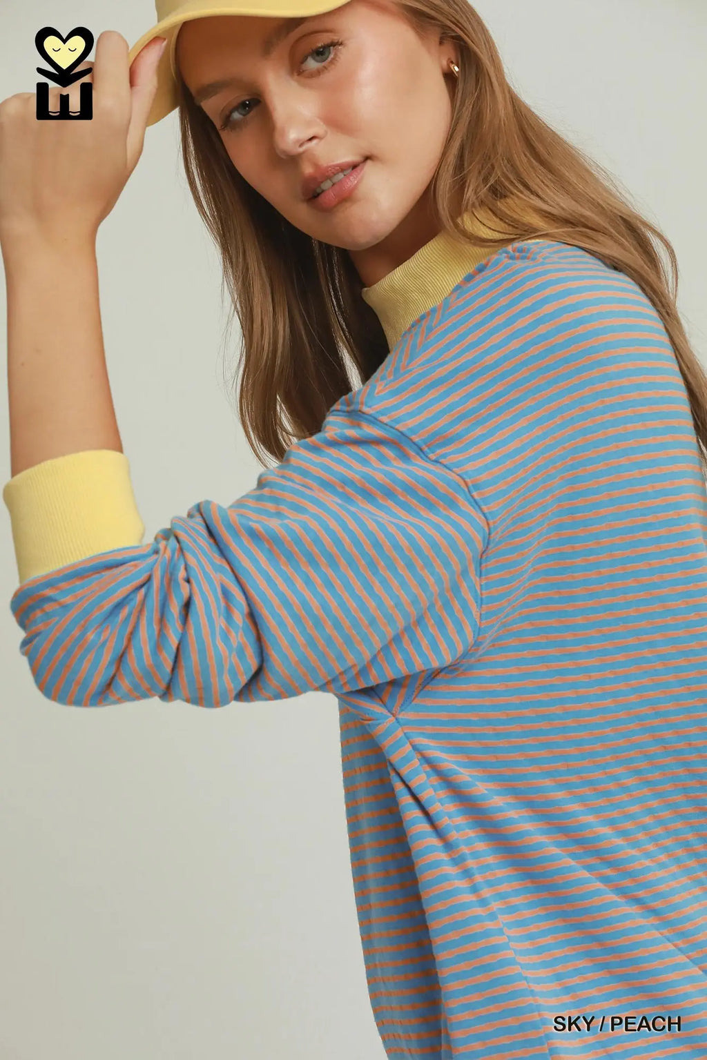 A woman wearing a light blue and orange striped long-sleeve top with a yellow mock neck collar, paired with a yellow hat. The photo is cropped from the chest up against a light gray background.