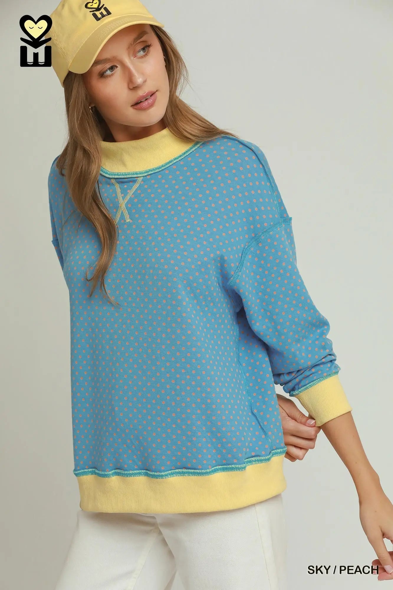 A woman wearing a light blue long-sleeve sweatshirt with a yellow dotted pattern, yellow ribbed collar, cuffs, and hem, paired with white pants and a yellow cap, against a light gray studio background.