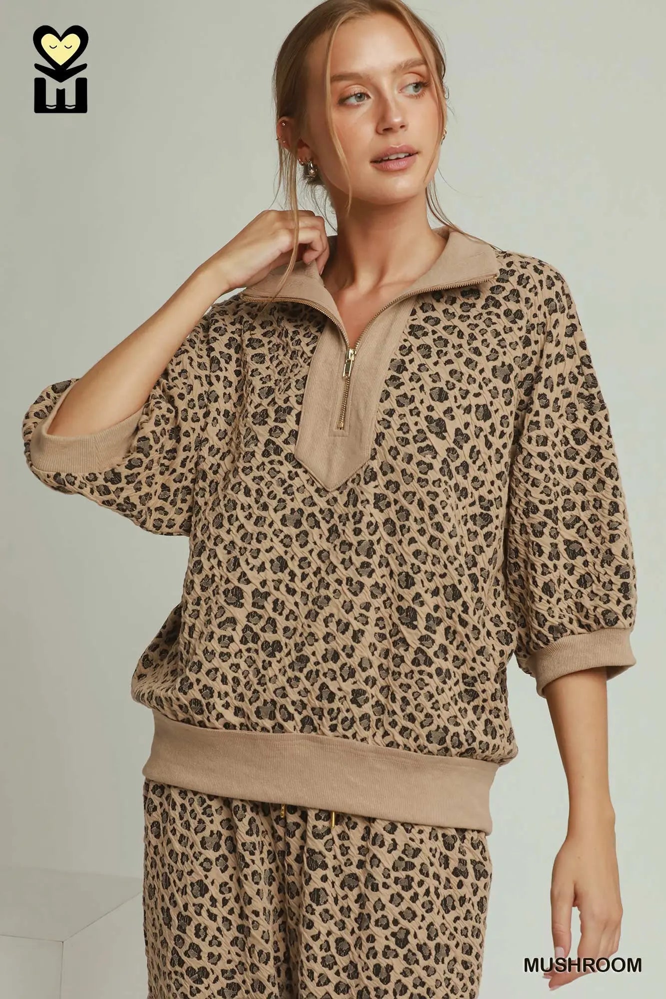 A woman modeling a matching two-piece lounge set in a beige and brown leopard print. The set includes a half-zip mock-neck sweatshirt and matching pants.