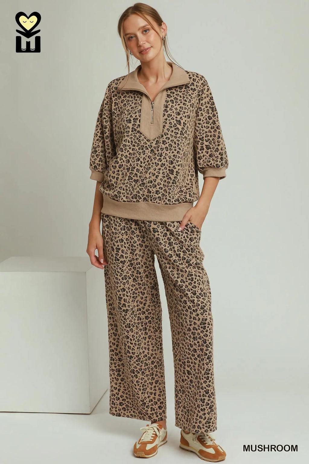 A woman modeling a matching two-piece lounge set with a brown and beige leopard print pattern, featuring a short-sleeve half-zip pullover and wide-leg pants, standing in a studio setting.