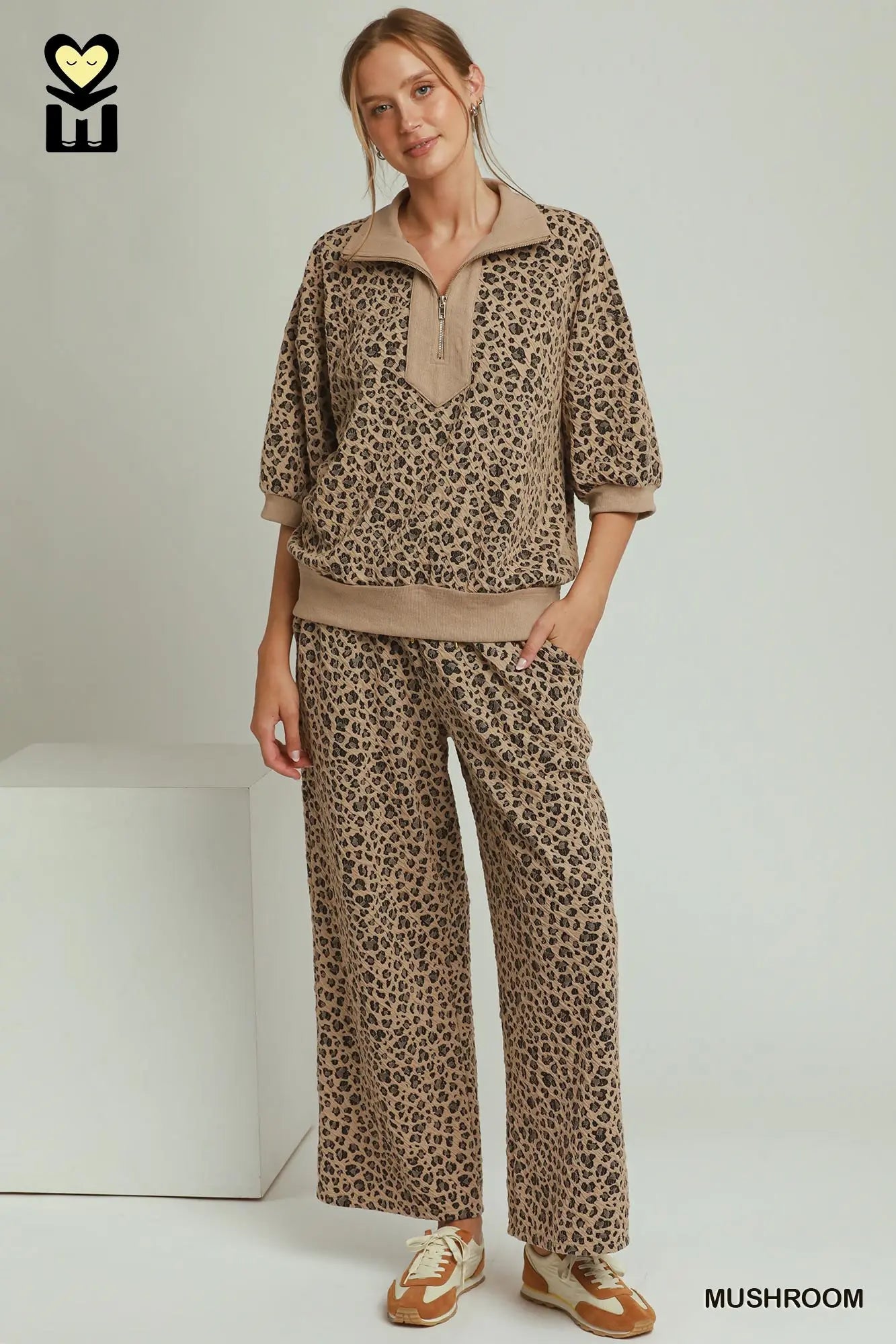 A woman modeling a matching two-piece lounge set with a brown and beige leopard print pattern, featuring a short-sleeve half-zip pullover and wide-leg pants, standing in a studio setting.