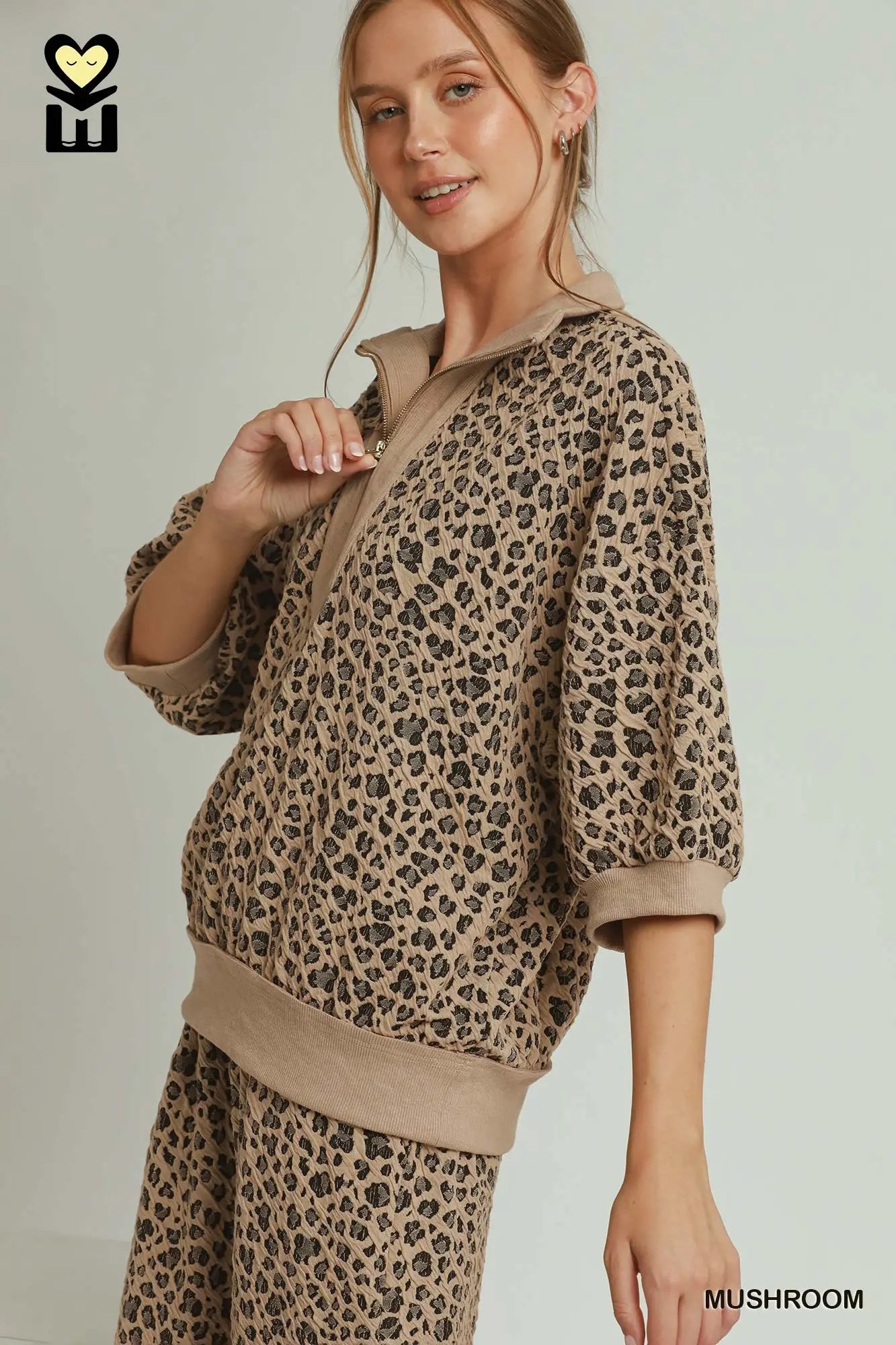 A woman modeling a matching beige and brown leopard-print lounge set, featuring a half-zip long-sleeve top and matching pants, against a plain light gray background.