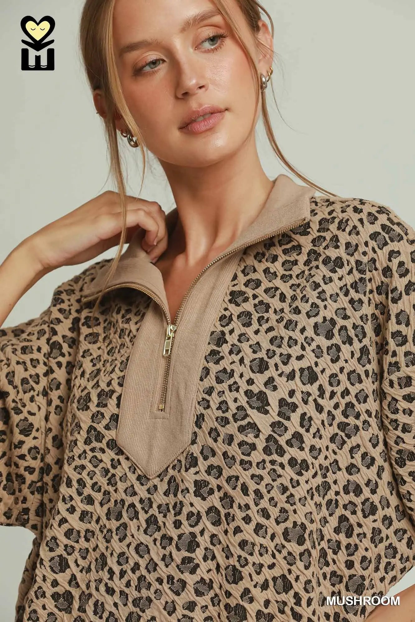 A woman wearing a beige and brown leopard-print half-zip pullover top with a solid beige collar, shown against a light gray studio background.