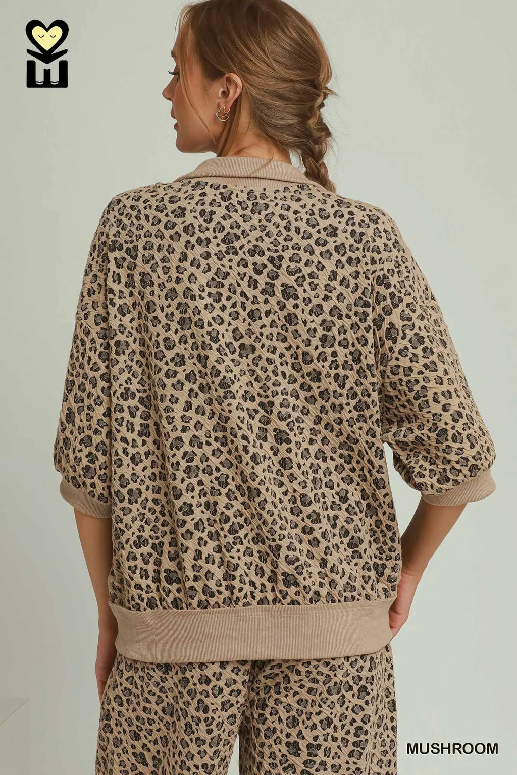 A woman shown from the back wearing a beige and brown leopard-print lounge set with a short-sleeve top and matching pants, against a plain light background.