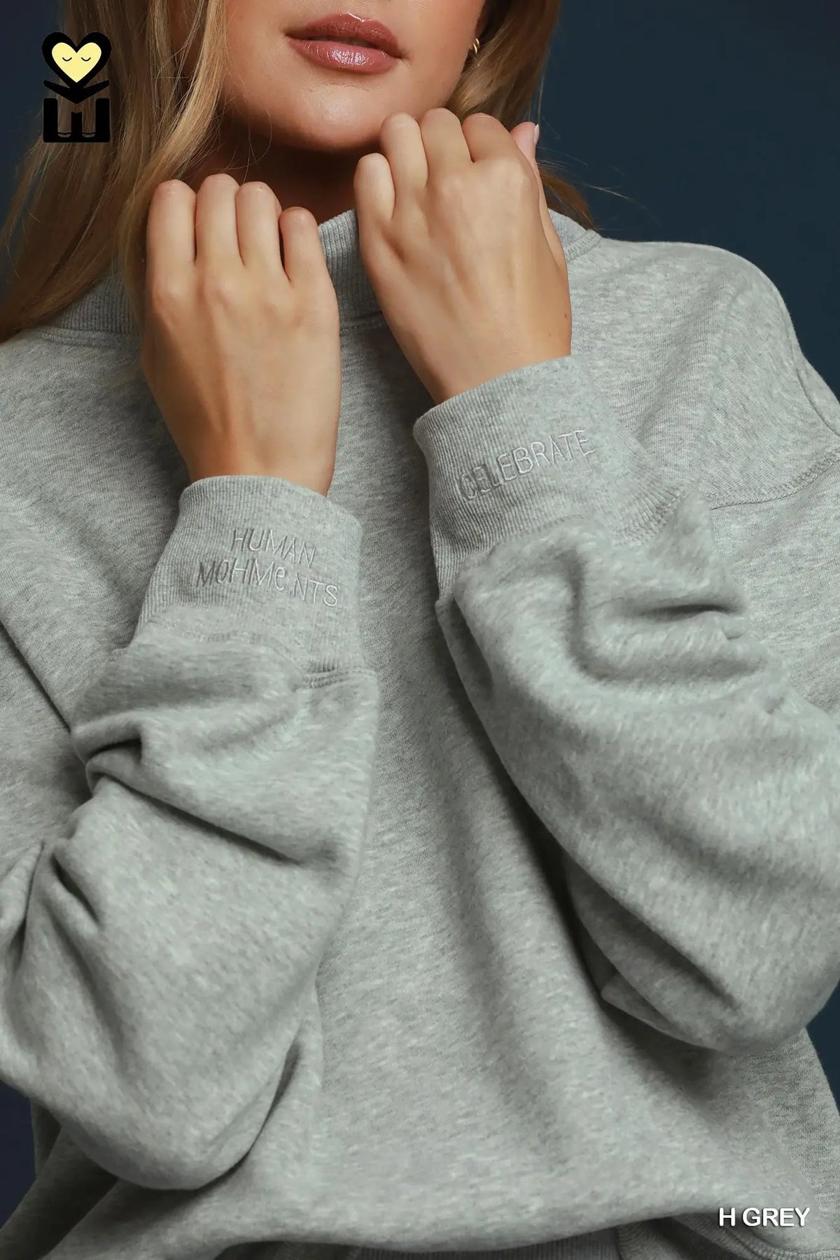 A close-up of a model wearing a light grey heather sweatshirt, showing the ribbed cuffs with tonal embroidered text and the soft texture of the fabric.