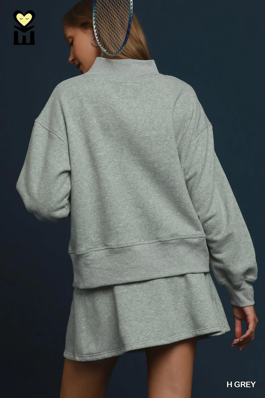 A woman shown from the back wearing a light heather grey sweatshirt and matching shorts, with a tennis racket hanging over her shoulder against a dark blue background.