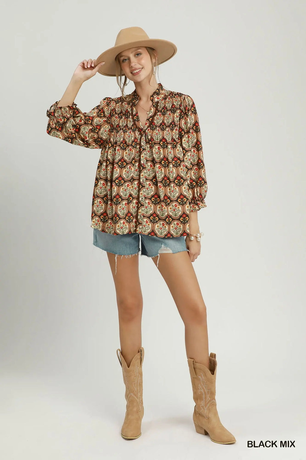 A woman modeling a bohemian-style long-sleeve button-up shirt with a brown, beige, and orange floral pattern, paired with denim shorts, tan cowboy boots, and a wide-brimmed tan hat against a light gray studio background.