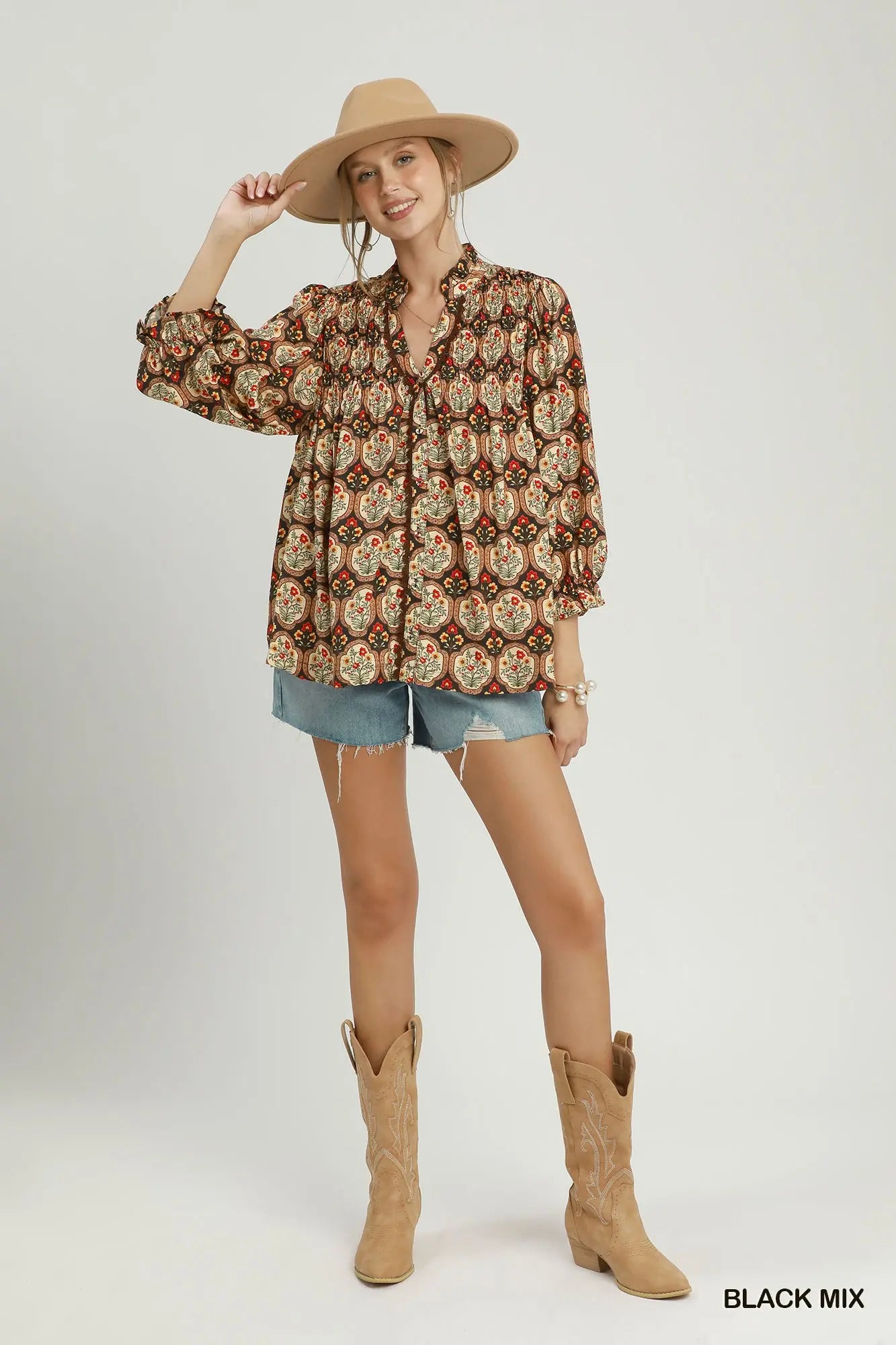 A woman modeling a bohemian-style long-sleeve button-up shirt with a brown, beige, and orange floral pattern, paired with denim shorts, tan cowboy boots, and a wide-brimmed tan hat against a light gray studio background.