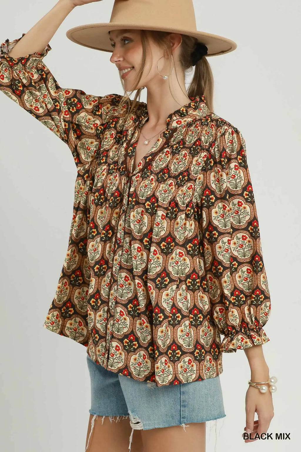 A woman wearing a bohemian-style long-sleeve blouse with a multicolor paisley and floral pattern on a brown base, paired with denim shorts and a wide-brimmed tan hat against a light gray studio background.