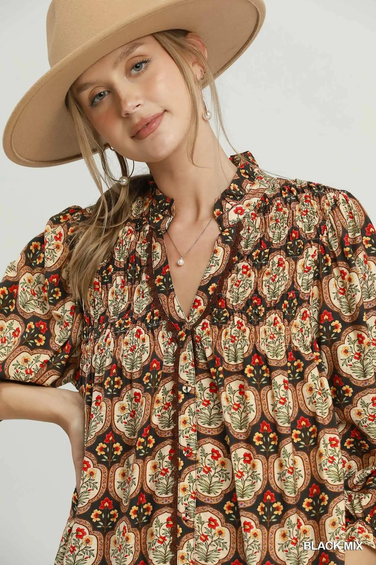 A woman wearing a beige wide-brim hat and a brown bohemian patterned blouse with red and yellow floral accents, photographed against a light gray studio background.