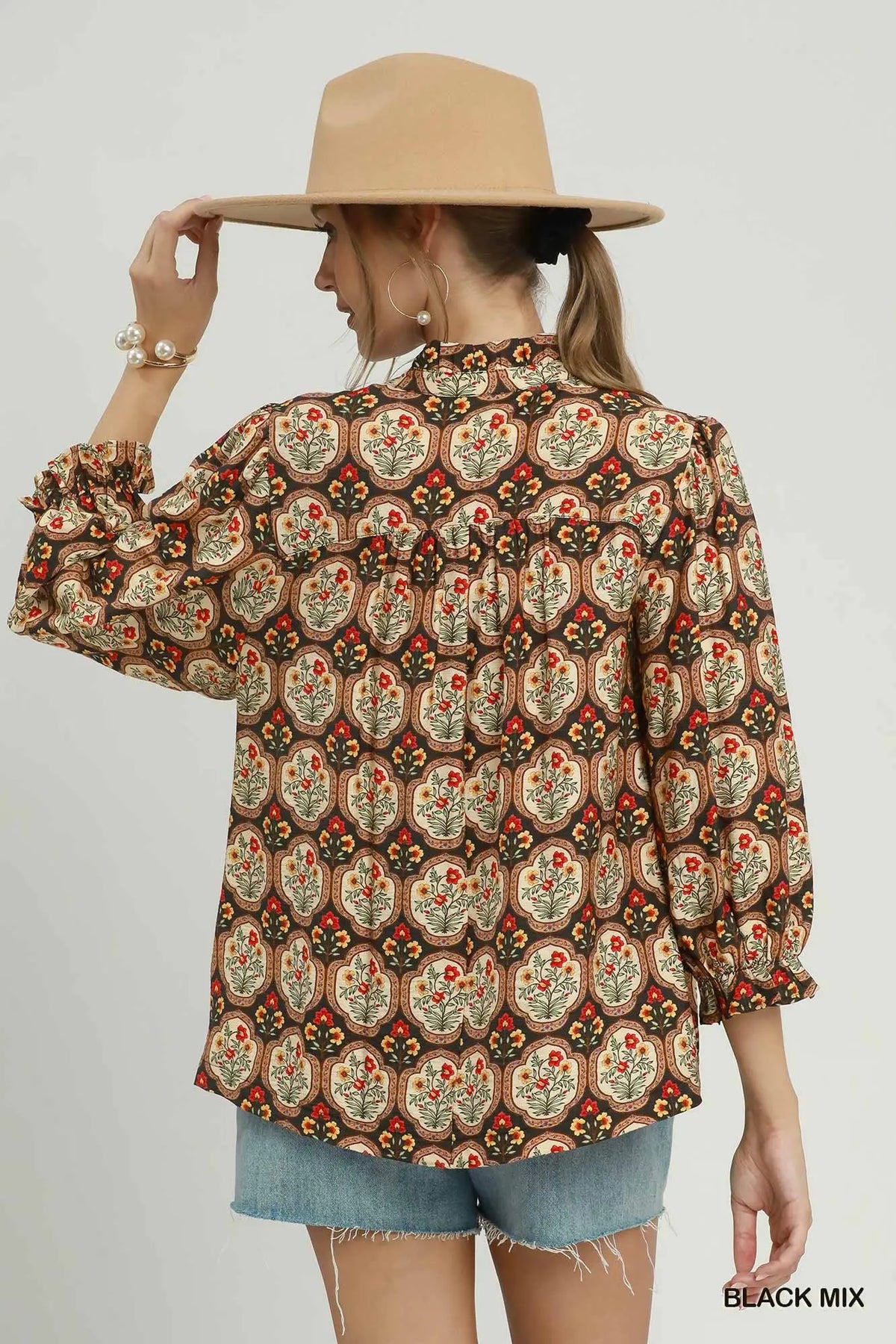 A woman wearing a bohemian-style patterned blouse with three-quarter ruffled sleeves, shown from the back. She is wearing a wide-brimmed tan hat and denim shorts against a light gray studio background.