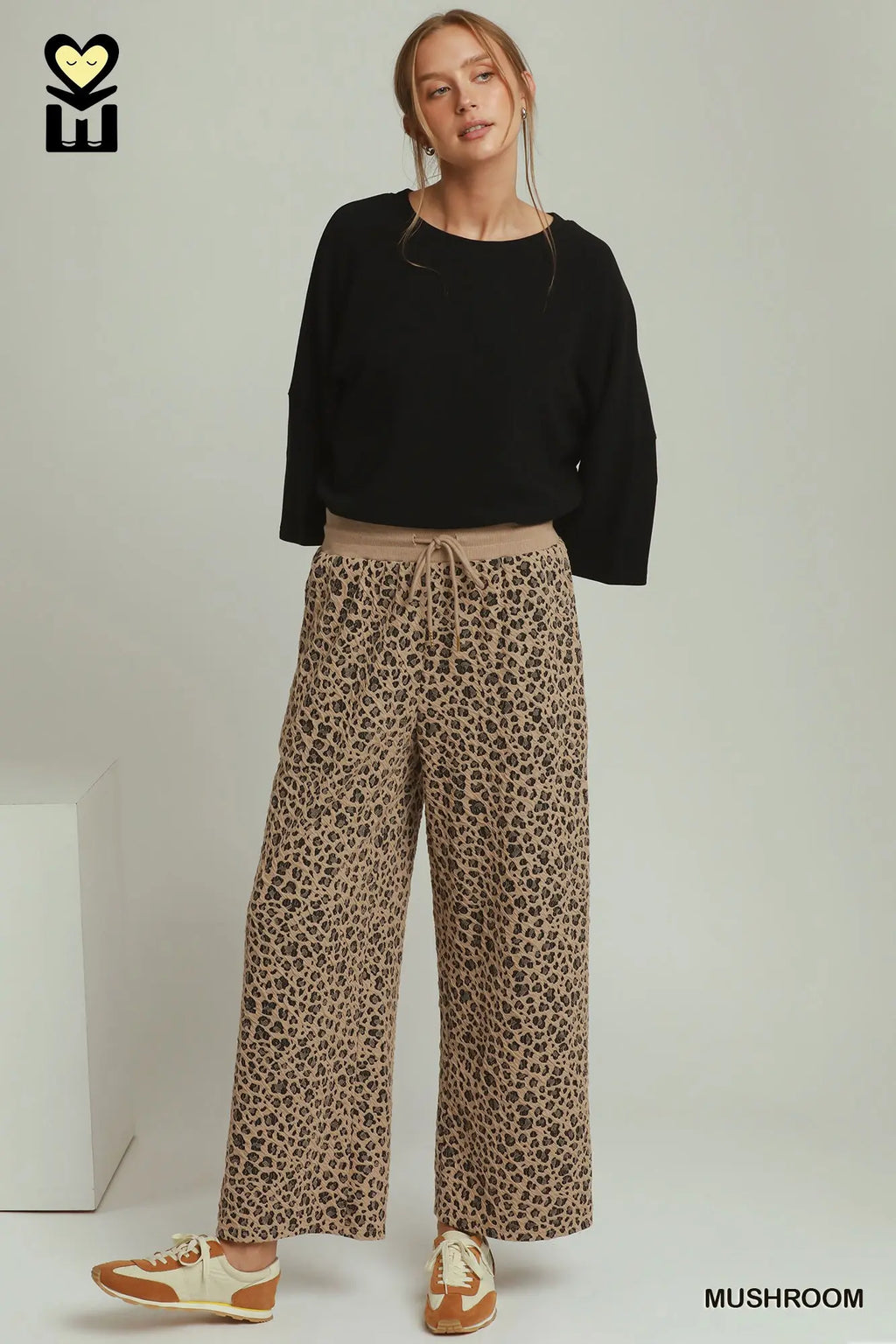 A model wearing wide-leg leopard print pants in a beige and brown shade, paired with a black long-sleeve top and beige-and-brown sneakers, standing against a light gray studio background.