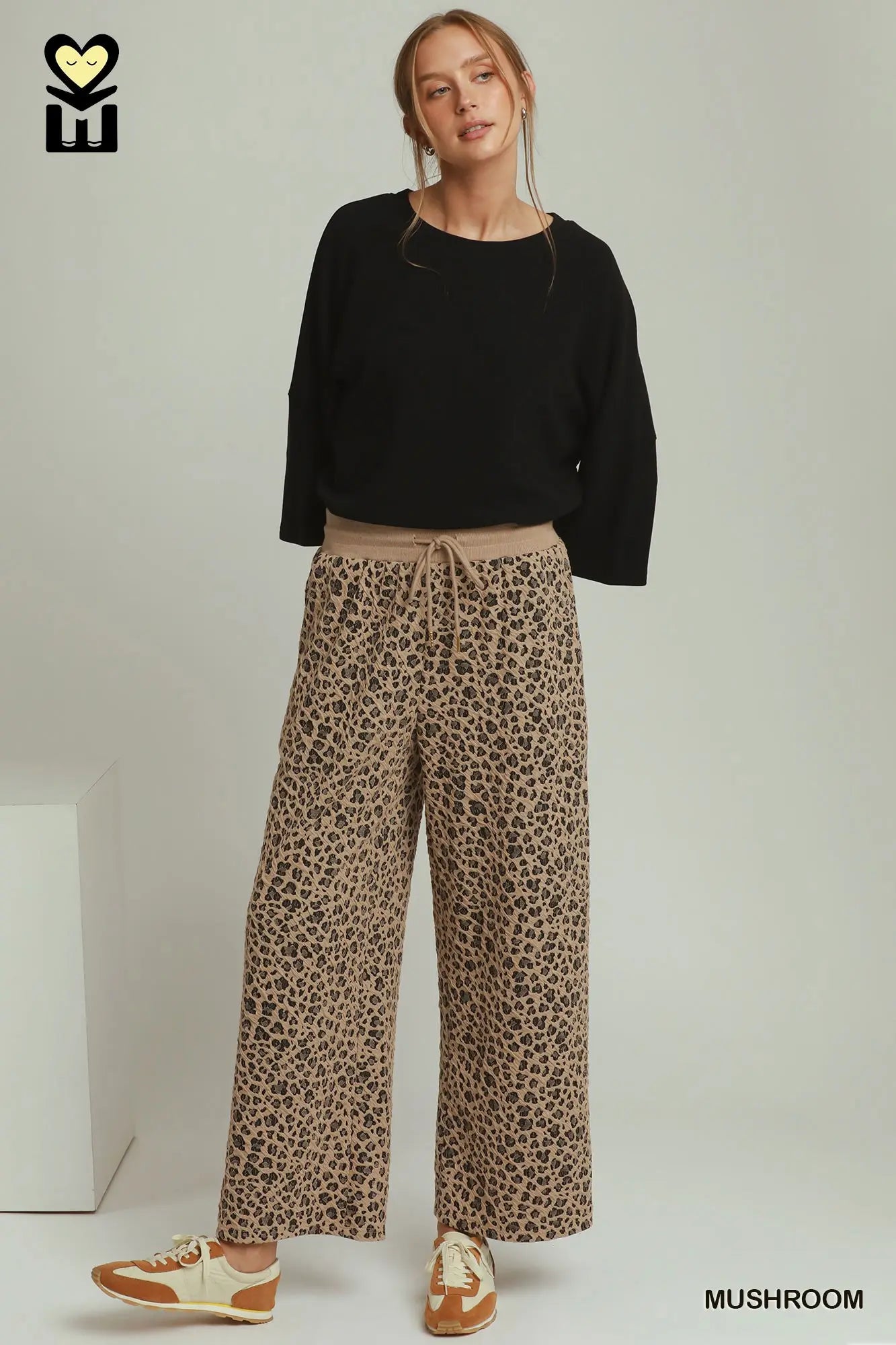 A model wearing wide-leg leopard print pants in a beige and brown shade, paired with a black long-sleeve top and beige-and-brown sneakers, standing against a light gray studio background.
