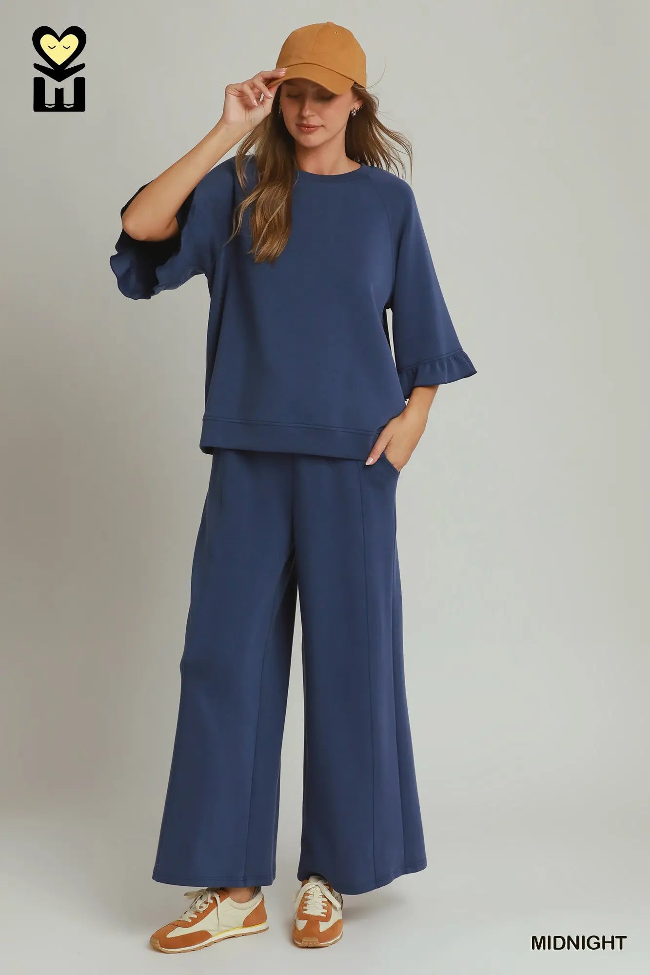 A woman modeling a navy blue two-piece lounge set consisting of a short-sleeve top with flared sleeves and matching wide-leg pants, styled with a tan cap, brown and cream sneakers, and a brand logo in the top left corner.