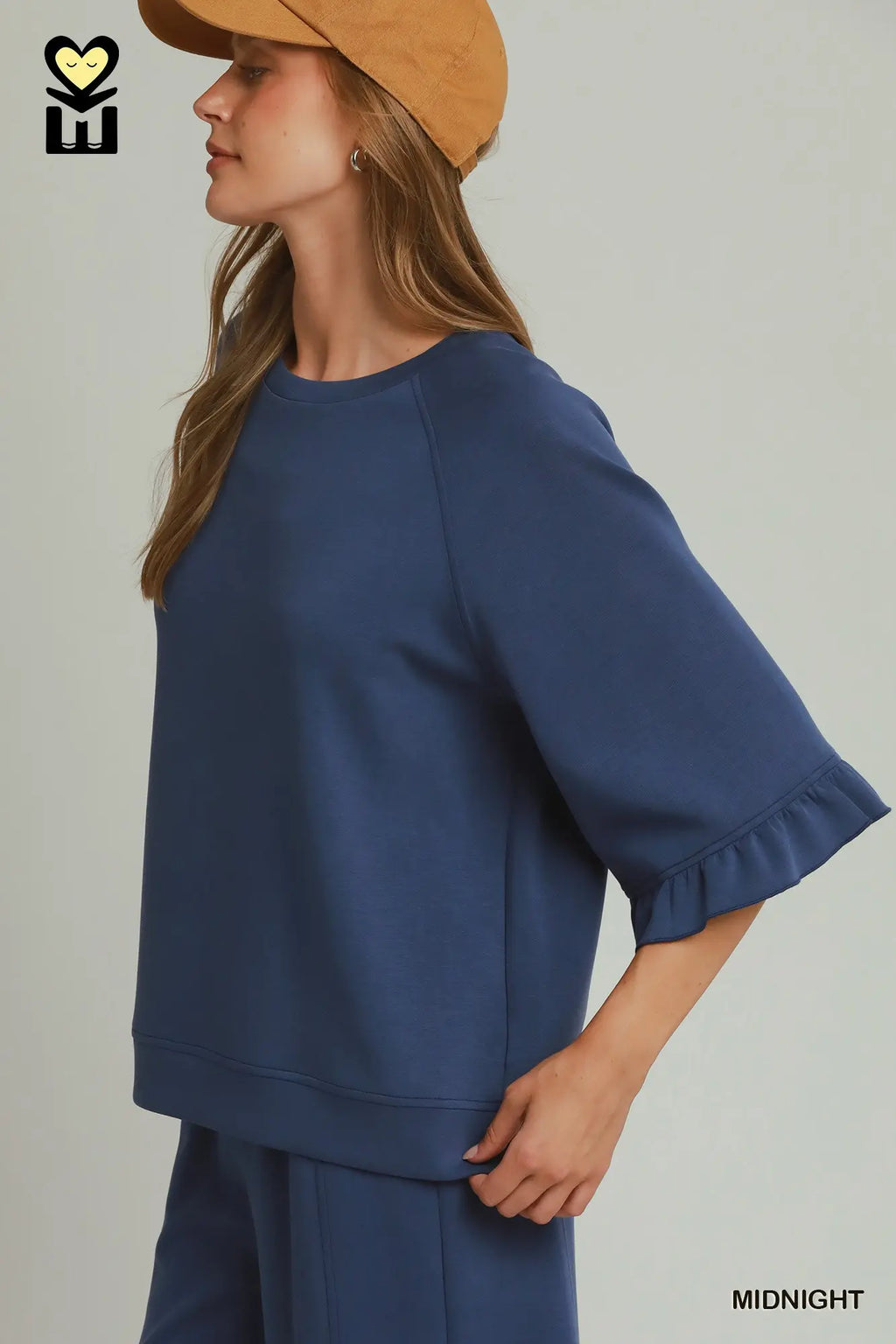 A woman wearing a navy blue short-sleeve top with ruffled cuffs and matching pants, paired with a tan hat, shown against a light gray studio background. The text 'MIDNIGHT' appears in the lower right corner.