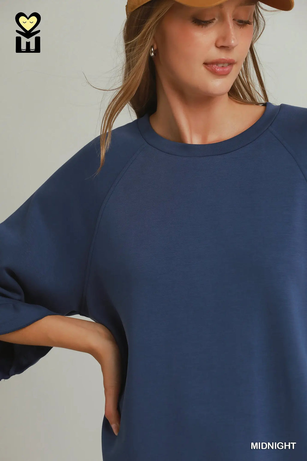 A woman wearing an oversized navy blue raglan sweatshirt, styled with a tan baseball cap. The image features a brand logo in the top-left corner and a color label 'MIDNIGHT' in the bottom-right corner.