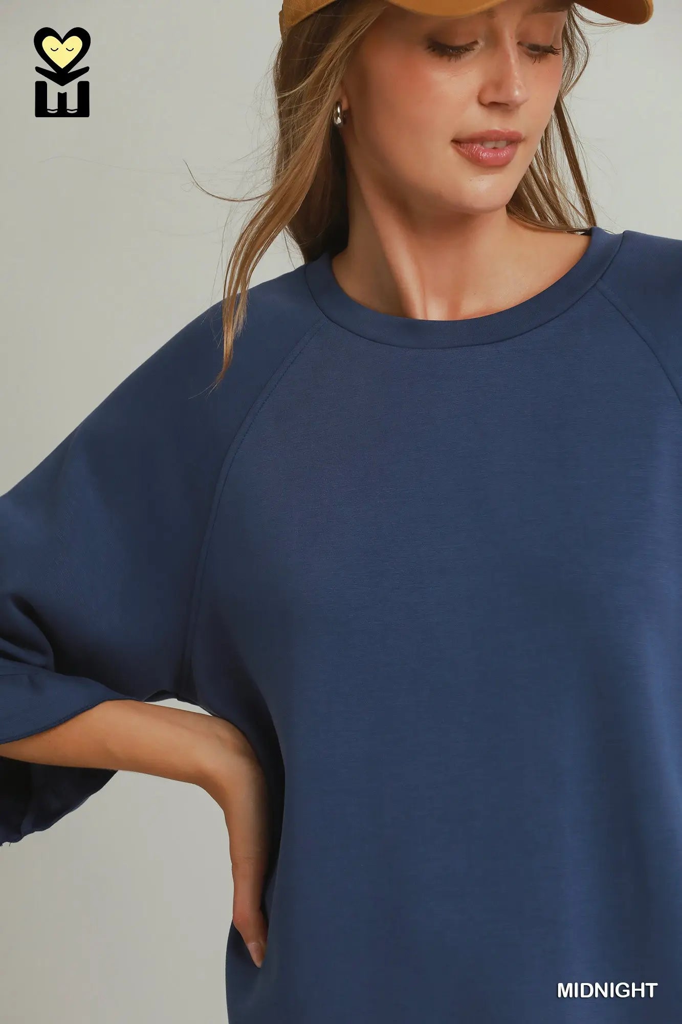 A woman wearing an oversized navy blue raglan sweatshirt, styled with a tan baseball cap. The image features a brand logo in the top-left corner and a color label 'MIDNIGHT' in the bottom-right corner.