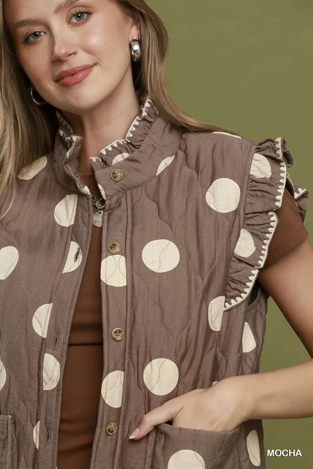 A woman wearing a brown sleeveless quilted vest with cream polka dots and ruffled trim, paired with a brown top, against an olive green background.