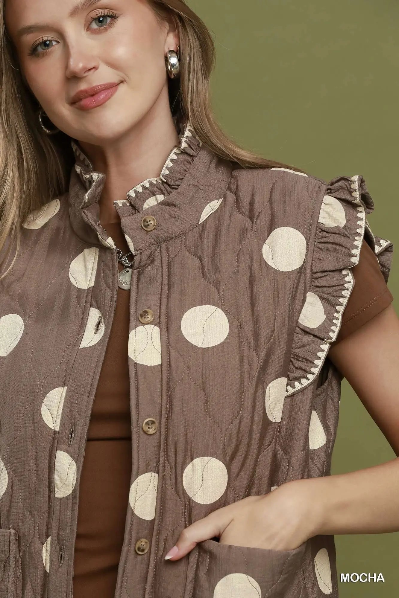 A woman wearing a brown sleeveless quilted vest with cream polka dots and ruffled trim, paired with a brown top, against an olive green background.