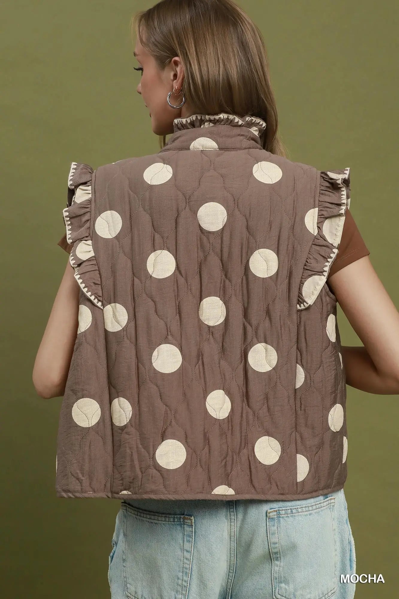 A woman shown from the back wearing a sleeveless quilted vest with a brown base and large cream polka dots, paired with light blue jeans against an olive green background.