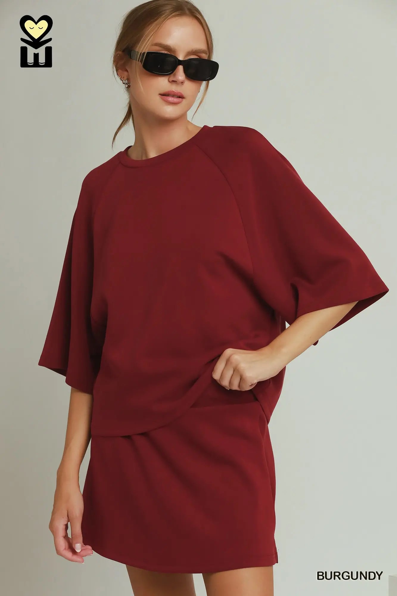 A woman modeling an oversized burgundy short-sleeve top and matching mini skirt set against a light gray studio background. She is wearing black sunglasses, and the brand logo 'HM' is visible in the top left corner.