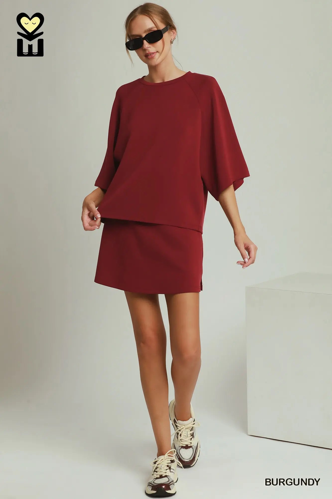 A woman modeling a burgundy two-piece set consisting of a short-sleeve top and a matching mini skirt, styled with beige sneakers and black sunglasses against a light gray studio background.