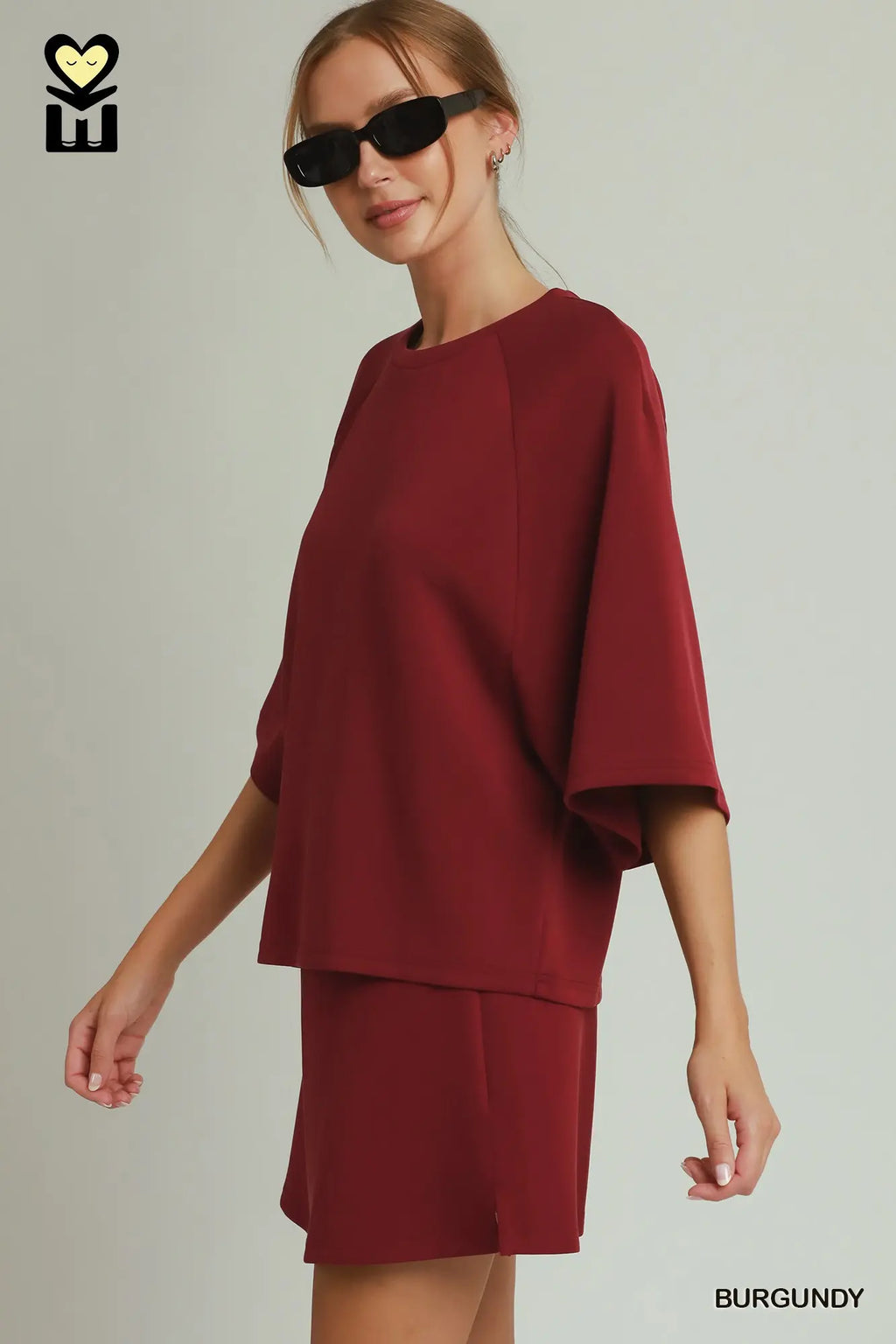 A woman modeling a burgundy two-piece set consisting of a loose, short-sleeved top and matching shorts, styled with black sunglasses against a light gray studio background.