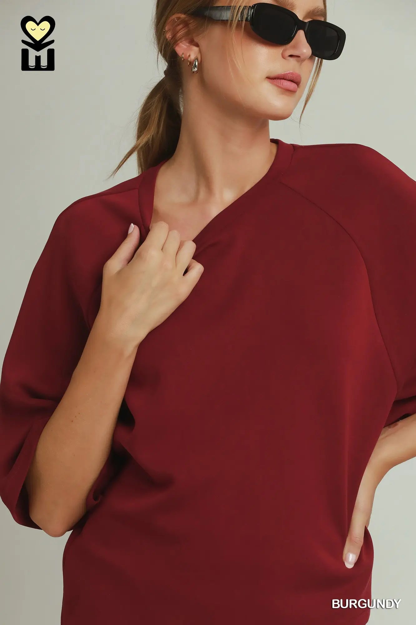 A woman wearing a burgundy V-neck short-sleeve top, styled with black sunglasses, against a light gray background. A brand logo is visible in the top-left corner, and the word 'BURGUNDY' is printed in the bottom-right corner.