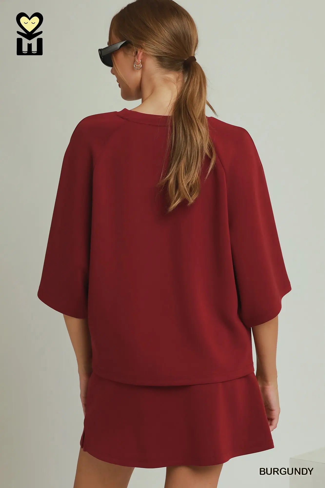 A woman shown from the back wearing a burgundy short-sleeve top and matching skirt set against a plain light gray studio background. The brand logo 'MH' appears in the top left, and the word 'BURGUNDY' appears in the bottom right.