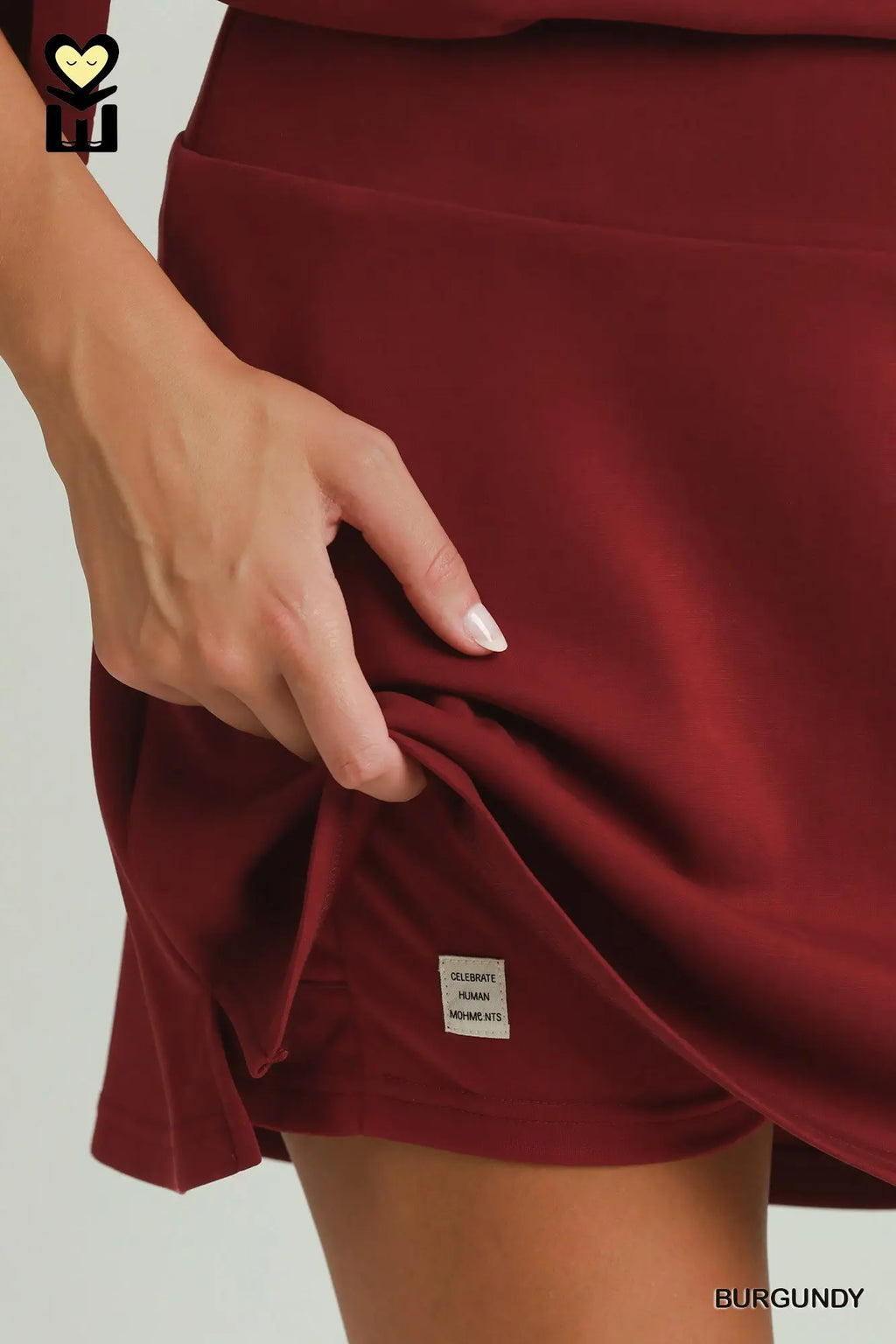 Close-up of a person wearing a burgundy athletic skirt, pulling the side hem to show a small white brand tag. The word 'BURGUNDY' appears in the bottom right corner.