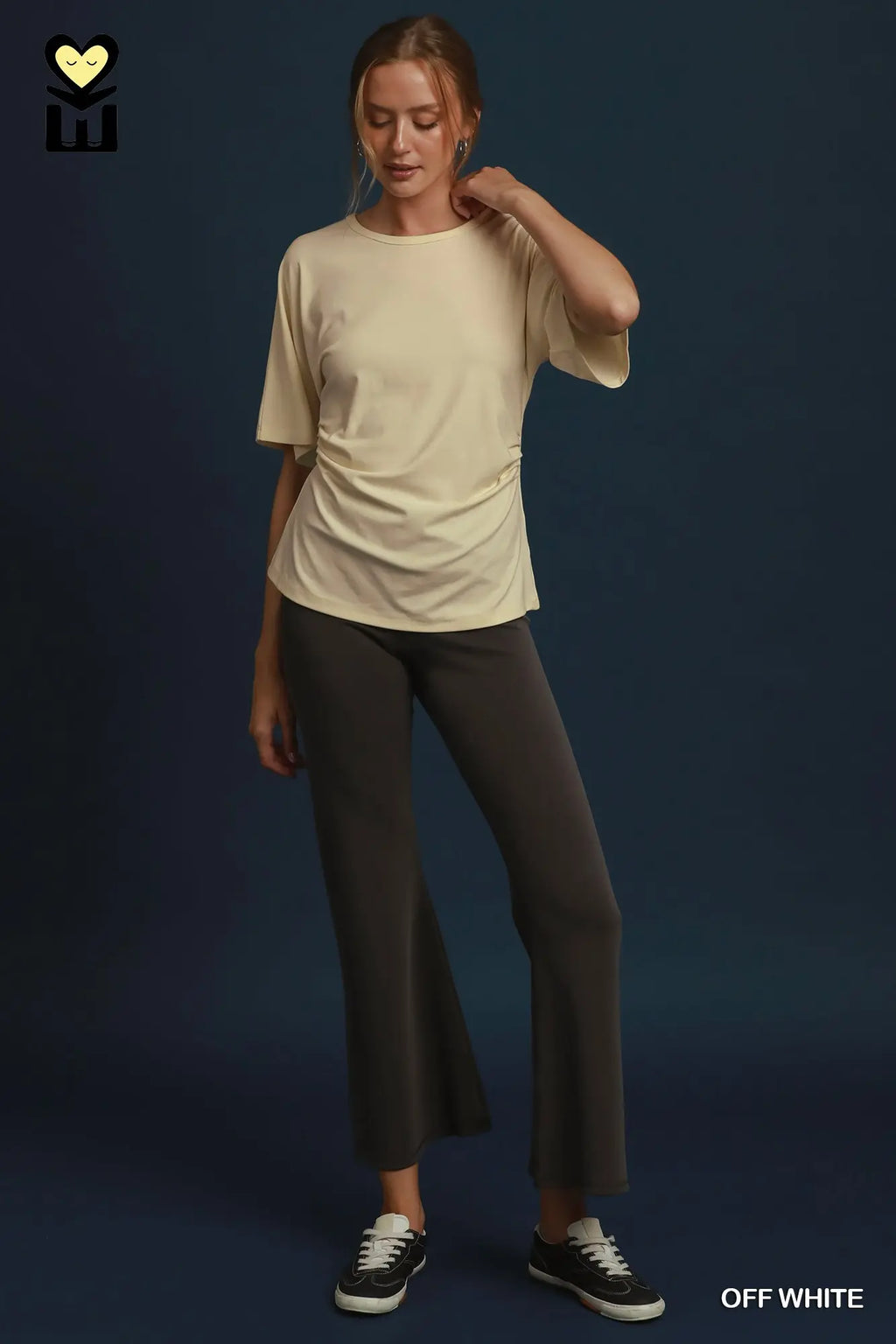 A woman modeling a short-sleeve crewneck t-shirt in off-white, styled with dark gray flared pants and black sneakers against a dark blue studio background.